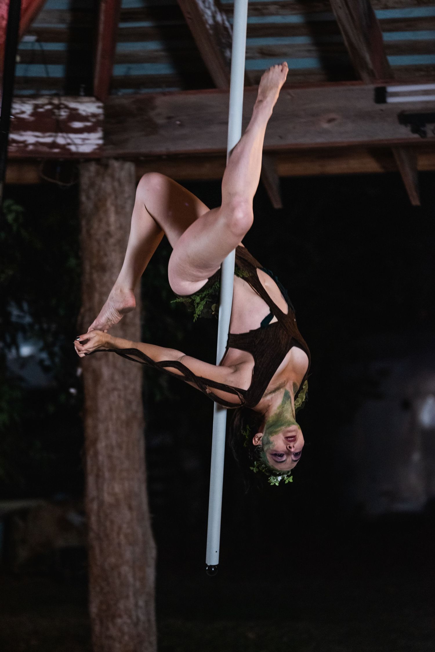 Aerial acrobat performs inverted pose on white pole against dark wooden beams.