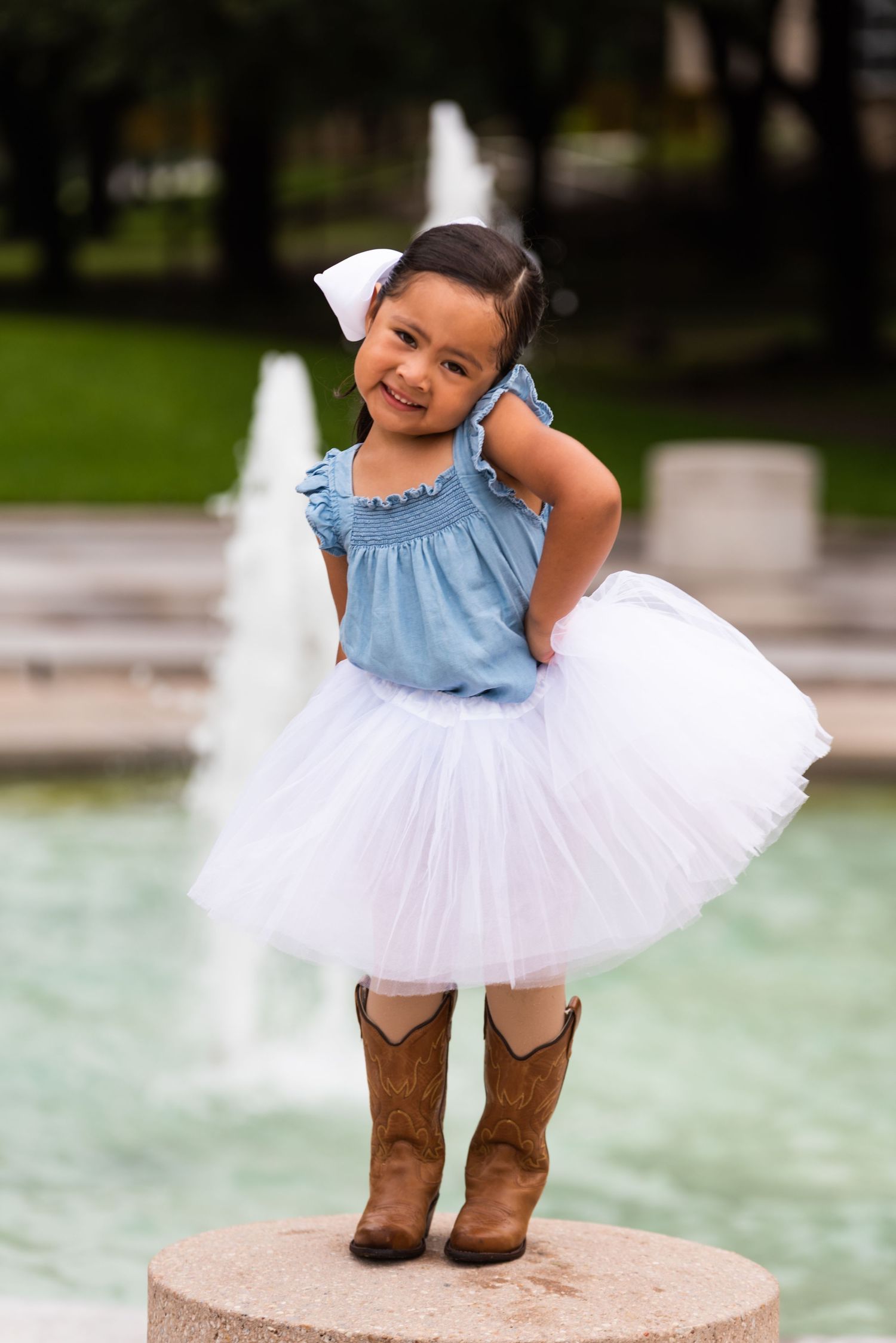 A young dancer poses by a fountain wearing a white tutu skirt, blue top and brown cowboy boots.