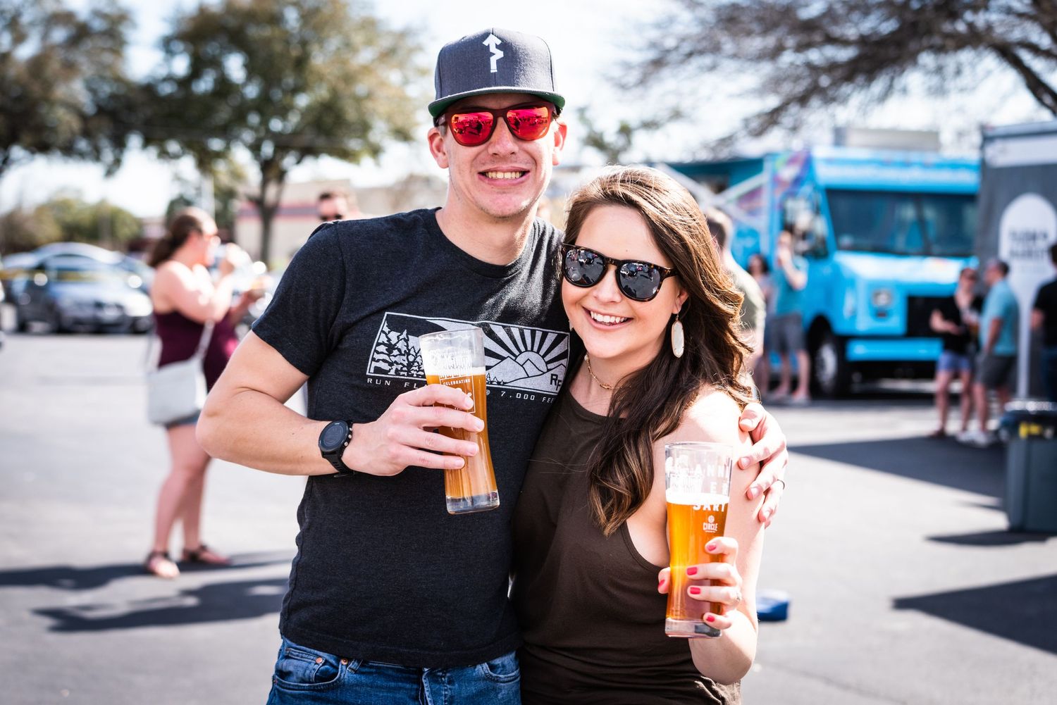 A couple enjoying beers at an outdoor festival on a sunny day.