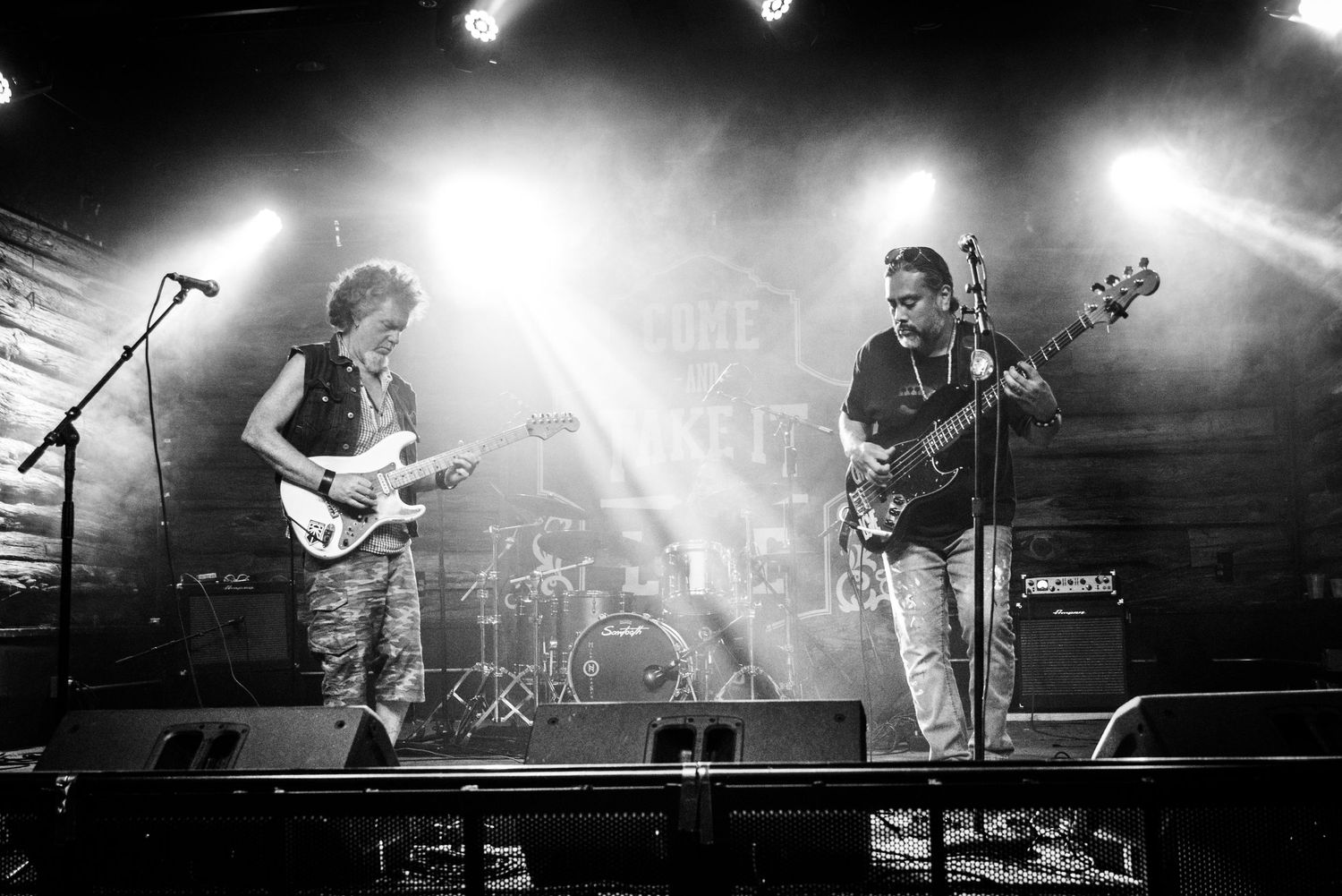 Two musicians perform together on stage with guitars and band equipment in black and white.
