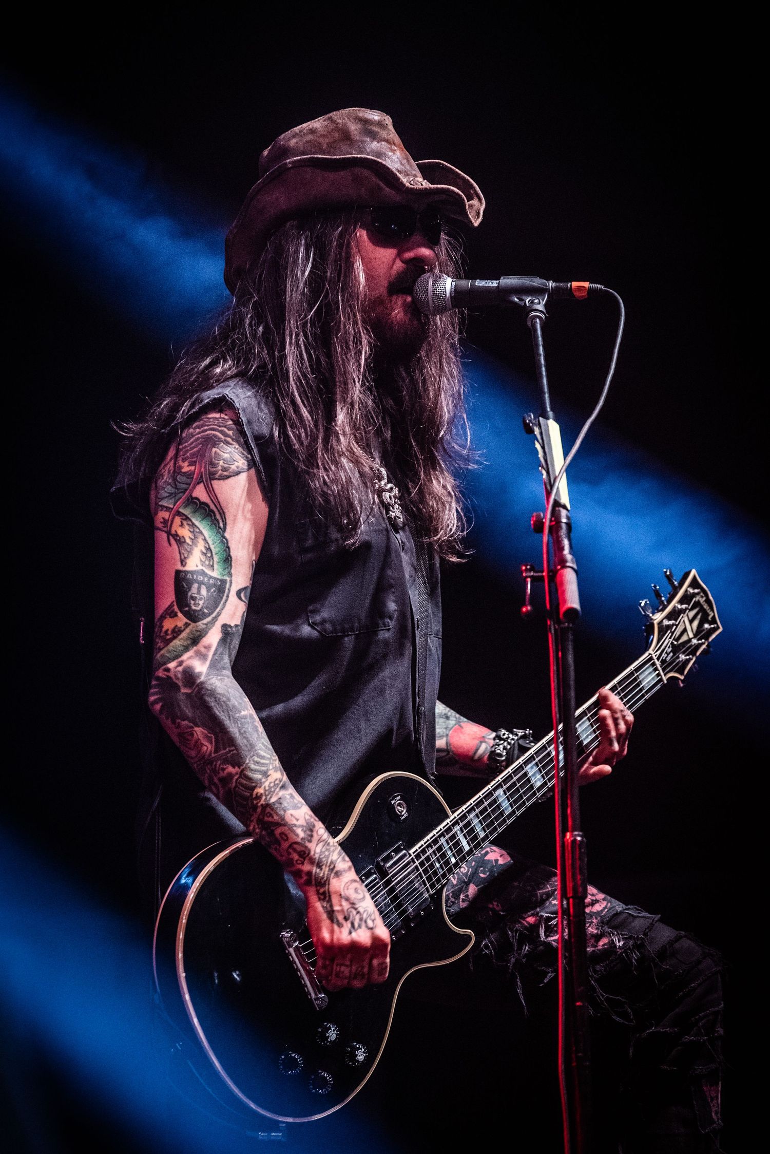 A tattooed musician plays guitar with long hair and a cap against a blue-lit backdrop.