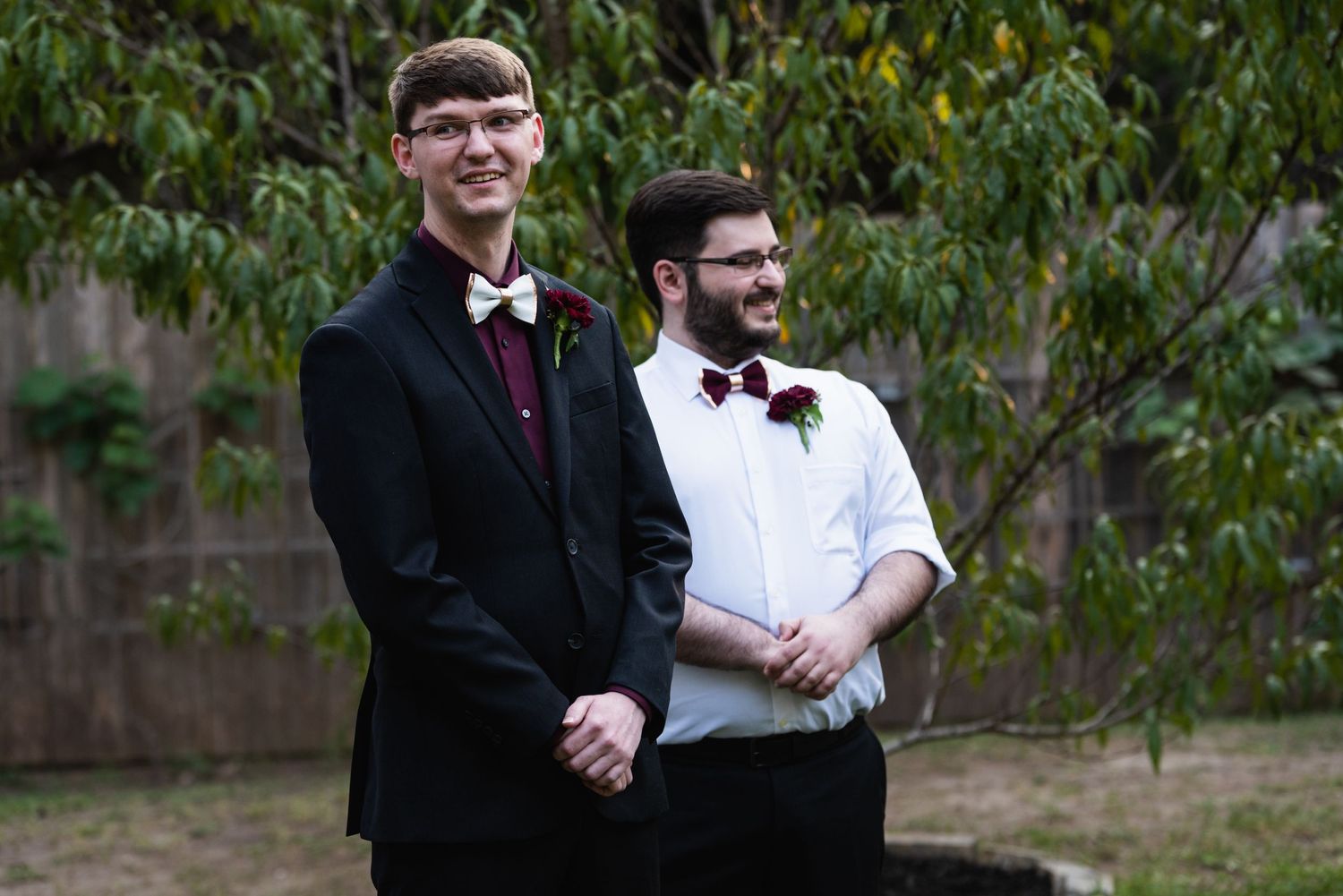 Two people in formal wedding attire stand together outdoors with burgundy bow ties.