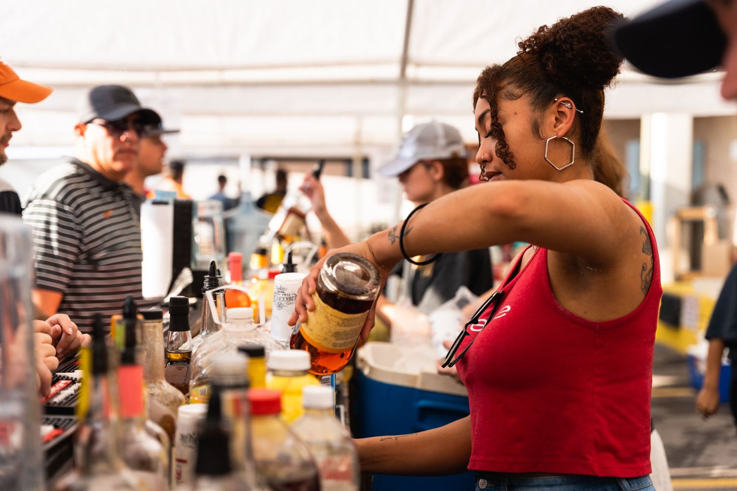 Bartender in red tank top mixes drinks at an outdoor event bar.