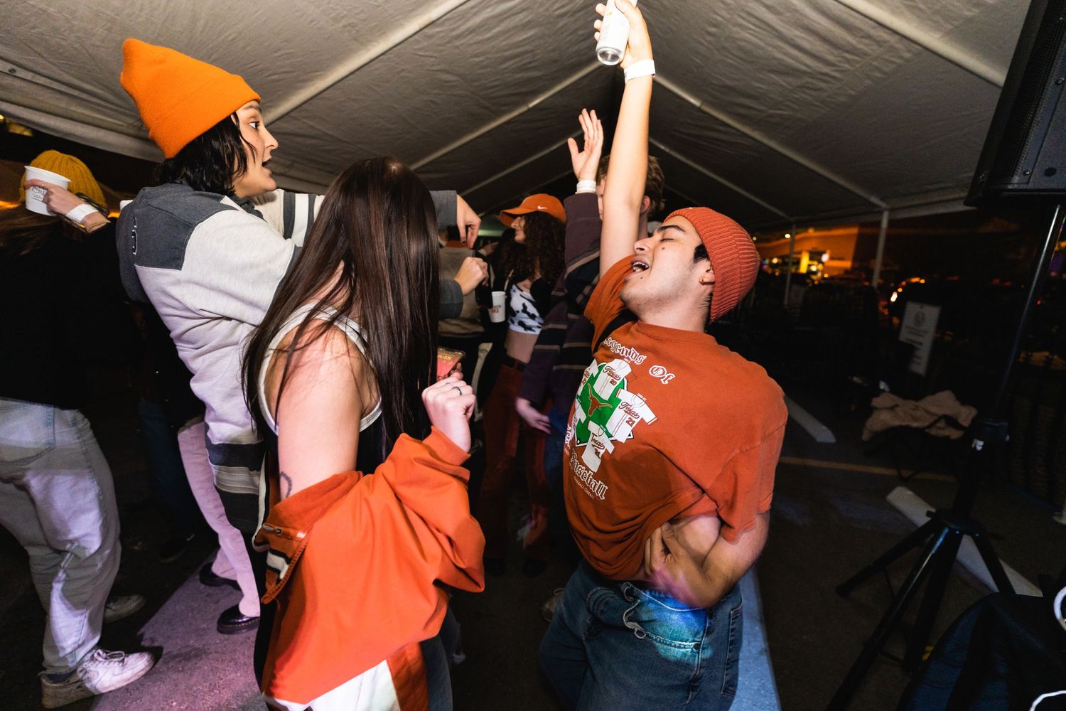 Partygoers dancing enthusiastically under a tent with one person wearing an orange beanie.