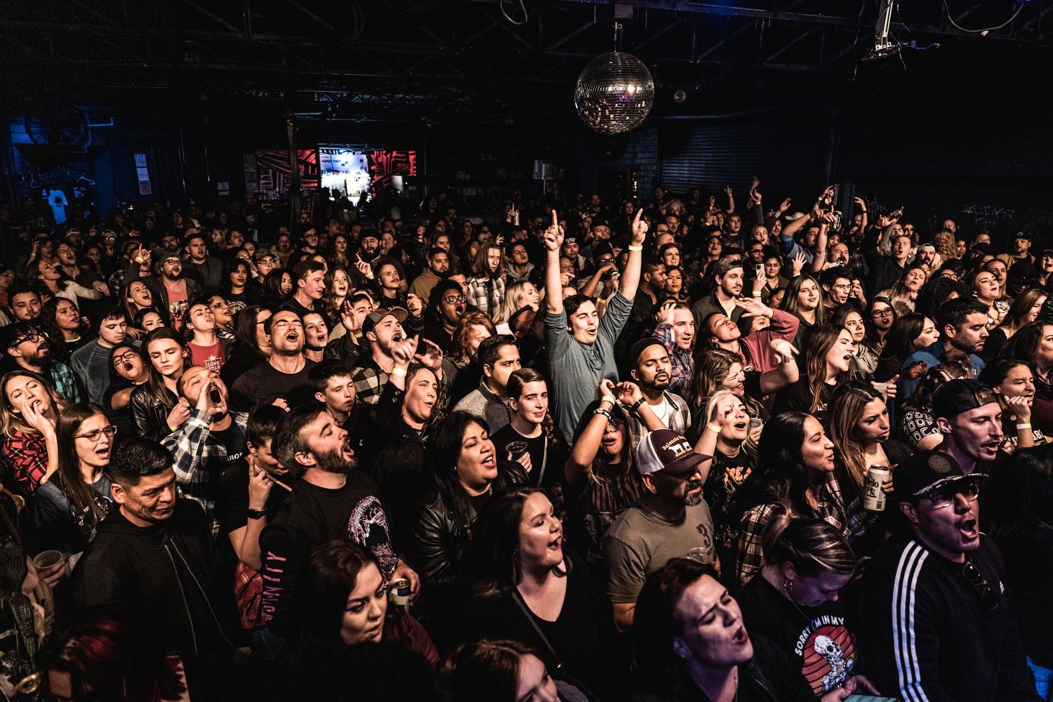 An energetic crowd fills a dark concert venue with raised hands and a disco ball overhead.