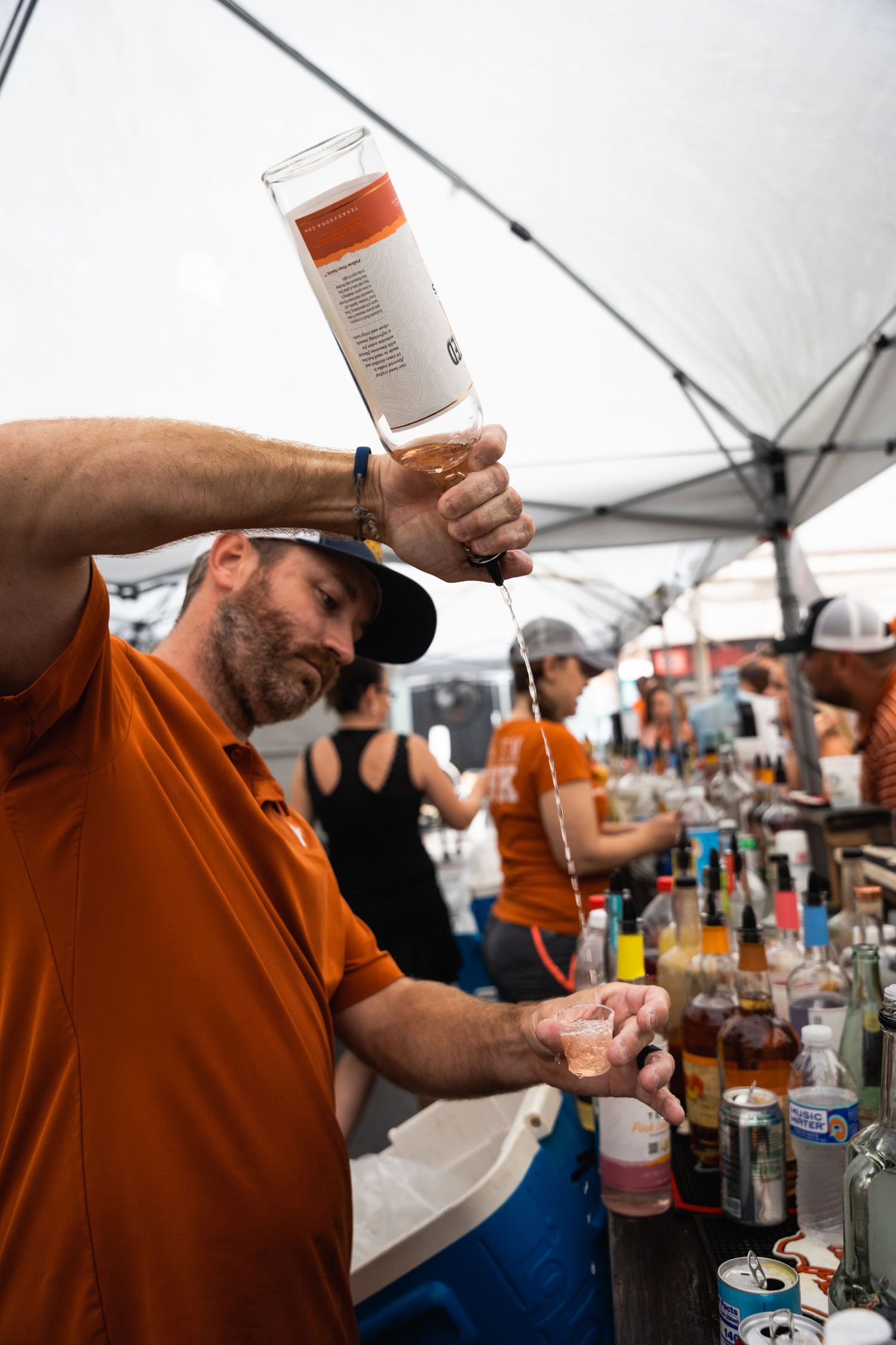 Bartender in orange shirt pours liquor at outdoor event bar setup with various bottles.