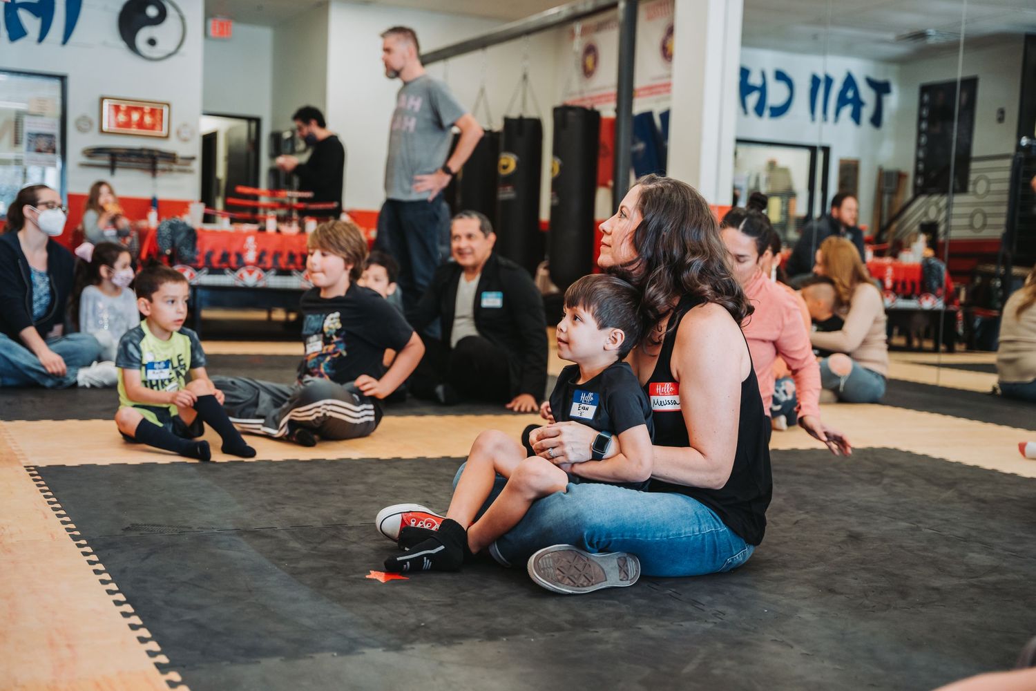 Children and adults sit on exercise mats during a martial arts class in a training studio.