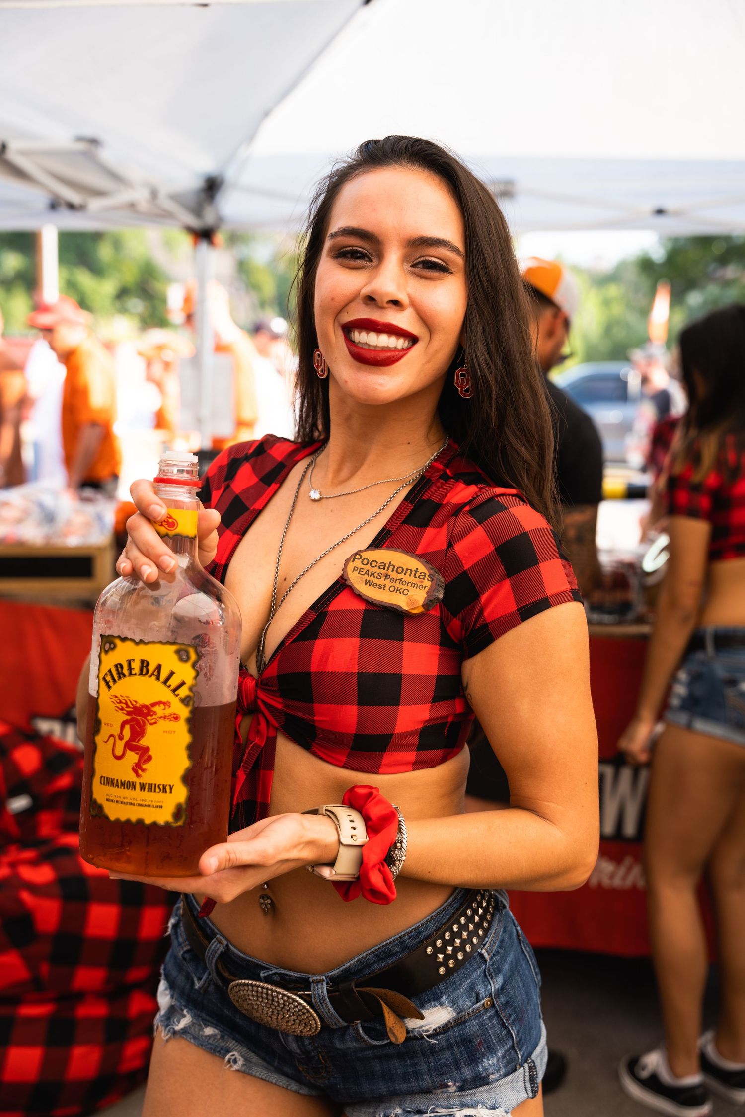 Woman in red plaid shirt holding a Fireball whiskey bottle at an outdoor festival event.