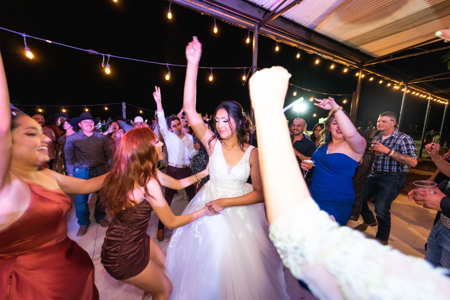 Guests dancing and celebrating with the bride at a lively wedding reception under string lights.