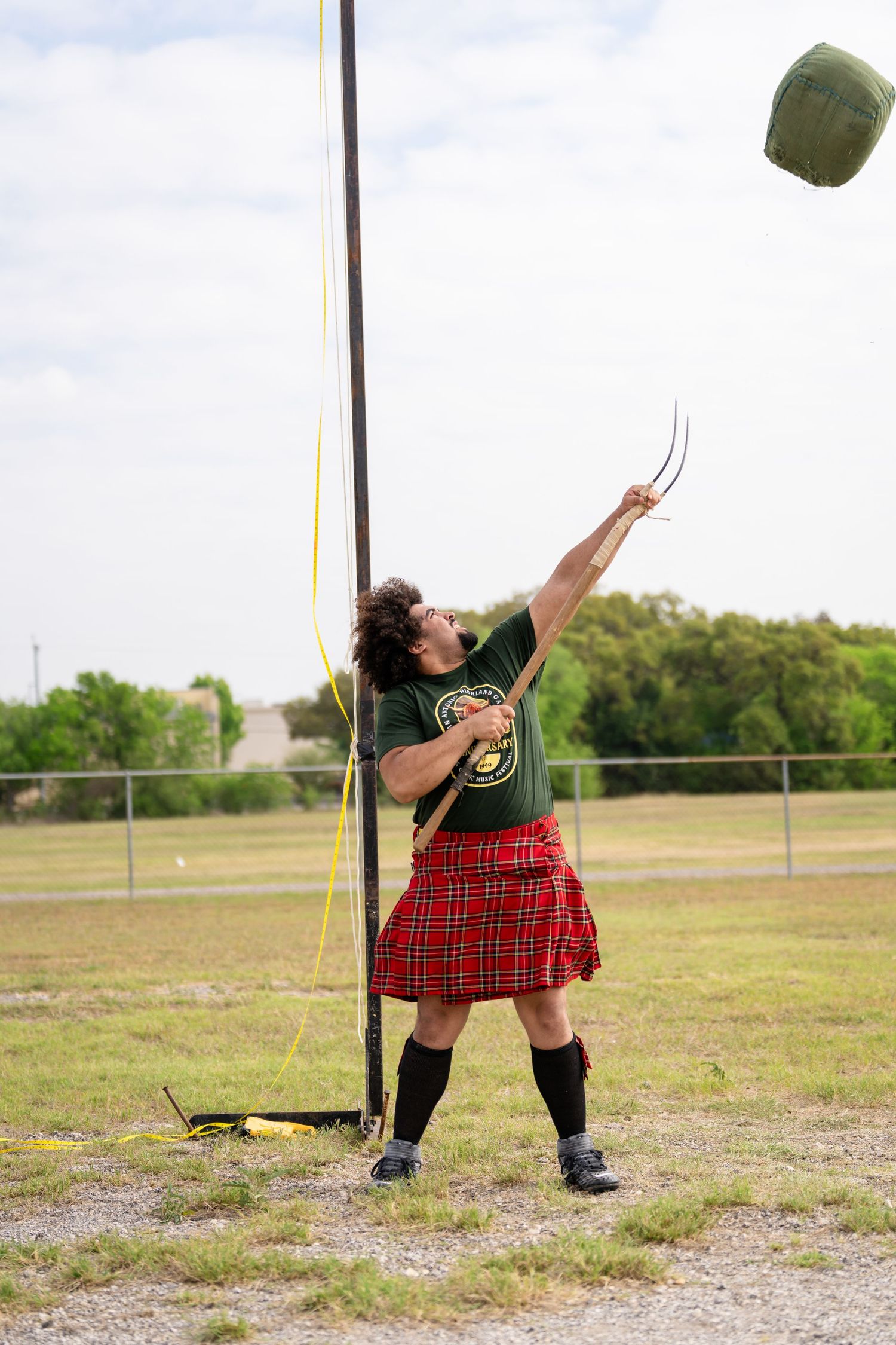 Man in red tartan kilt throwing weighted bag at Highland Games competition outdoors.