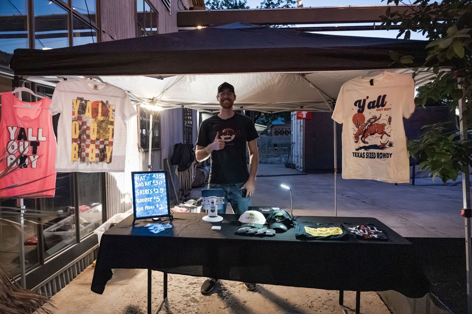 Man selling merchandise at an outdoor market booth with t-shirts displayed under a canopy at night.