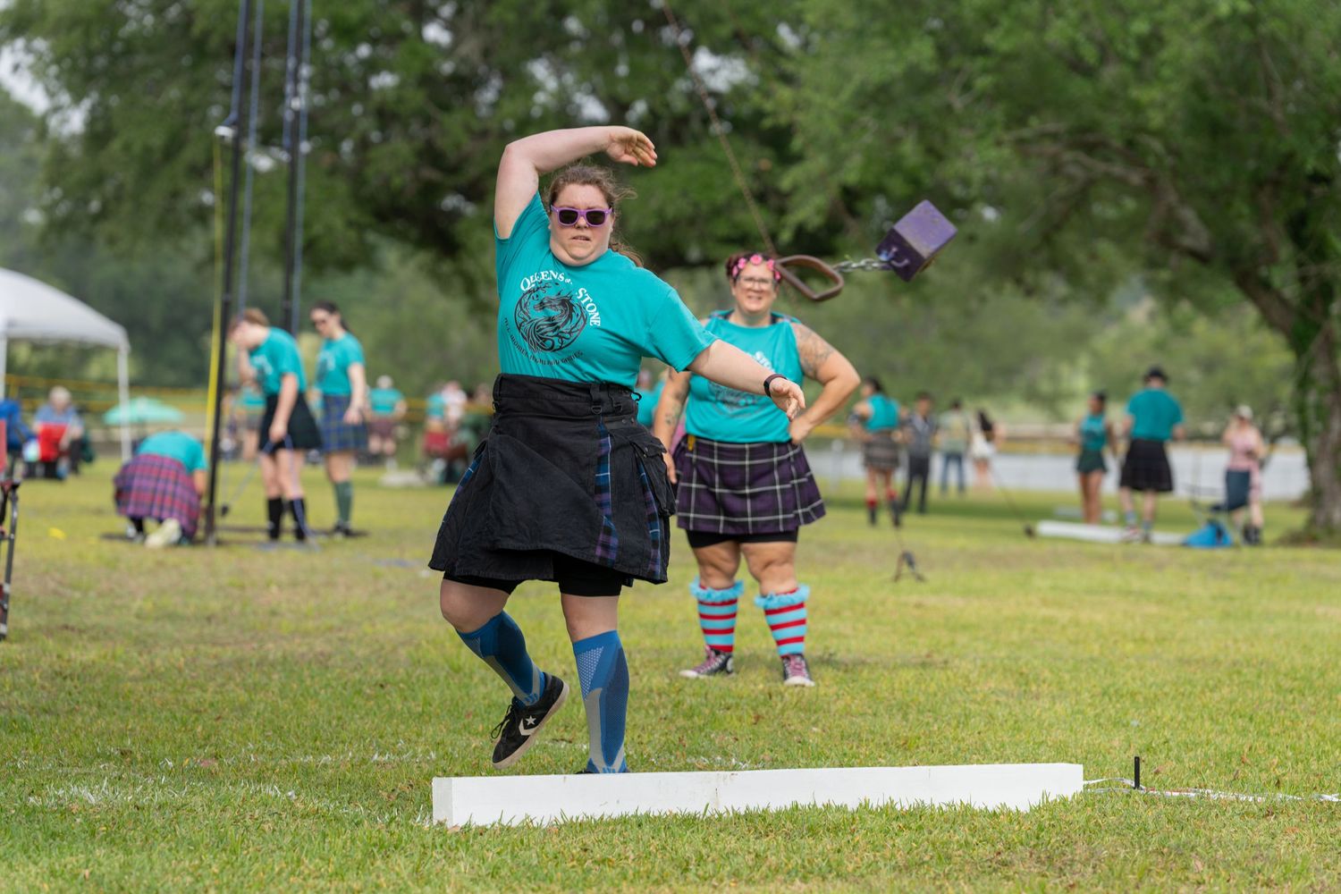 Women competing in Highland Games athletics, wearing teal shirts and kilts, throwing on a grass field outdoors.
