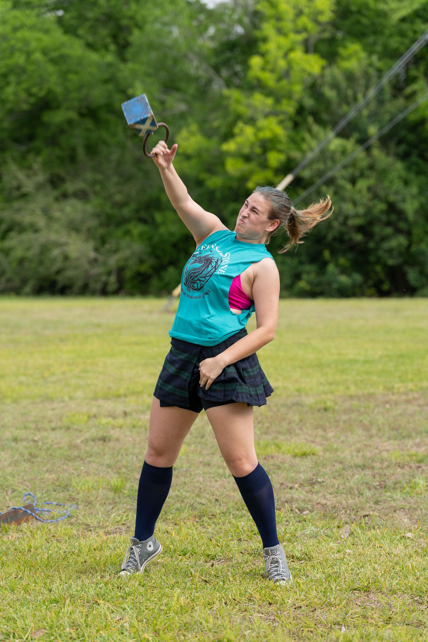 Woman in tartan skirt and knee socks throws weight over bar at Scottish Highland Games competition.