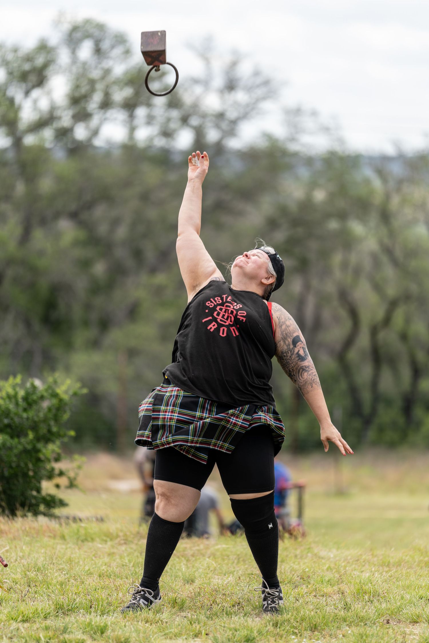 Athlete in kilt competes in Highland Games weight toss, reaching skyward on grassy field.