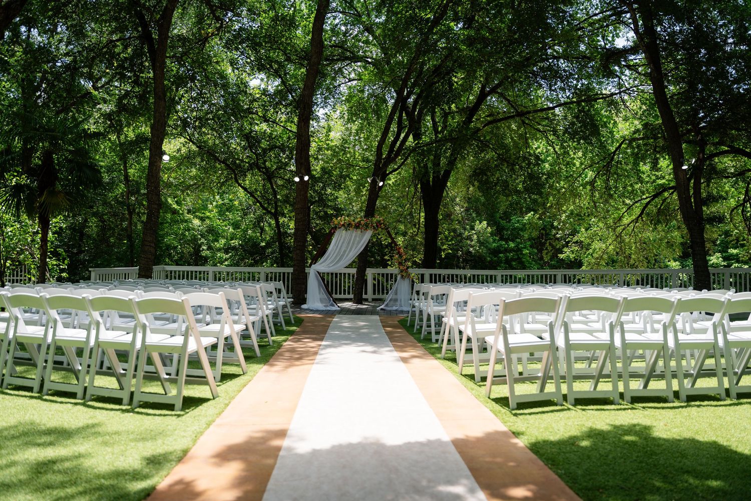 Outdoor wedding ceremony setup with white folding chairs, a rustic aisle runner, and a floral arch surrounded by lush green trees.
