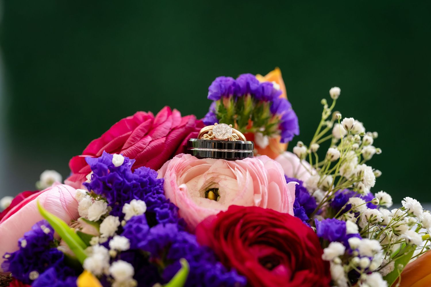 Wedding rings resting on a vibrant bridal bouquet of red, pink, purple flowers and baby's breath.