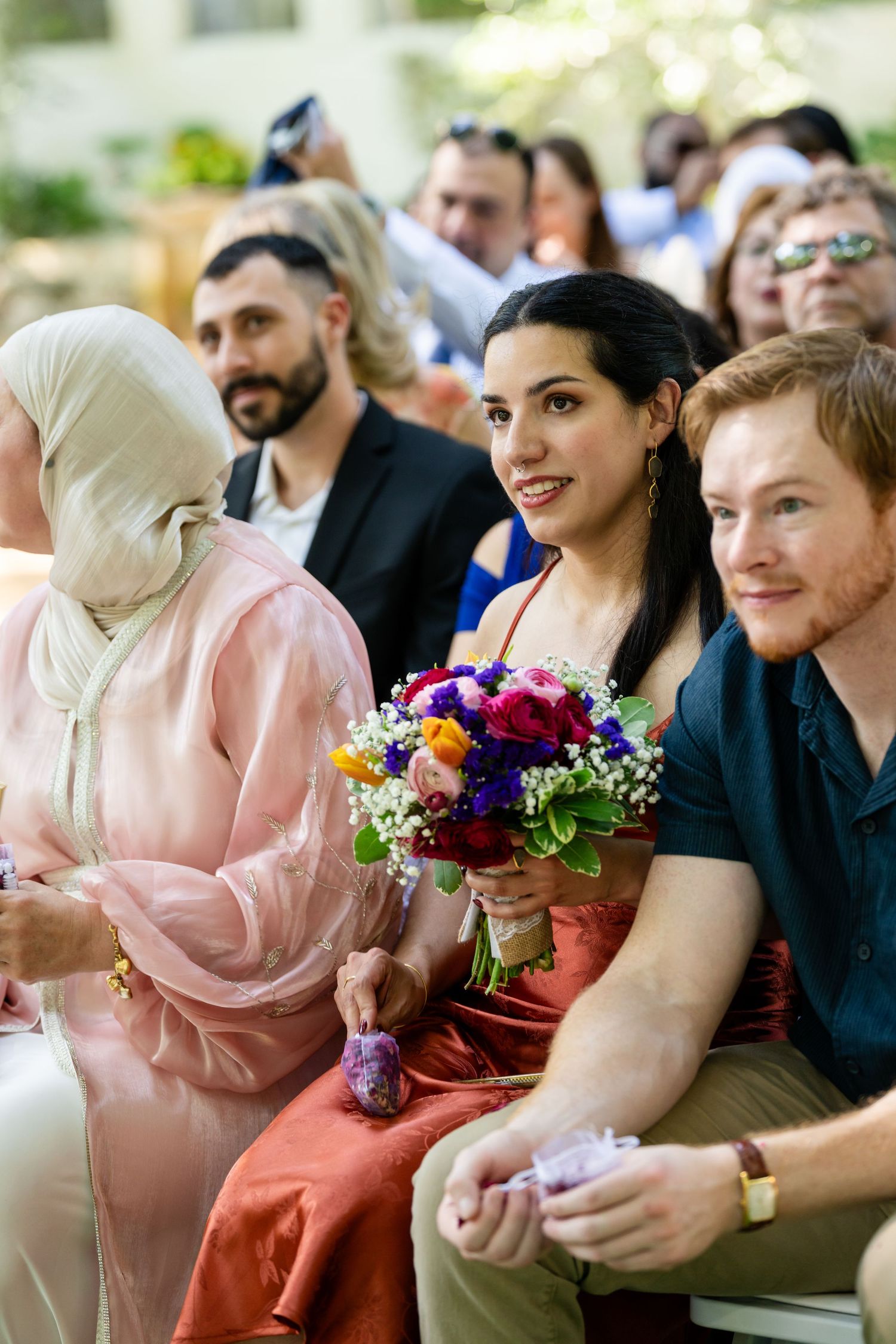Diverse wedding guests seated outdoors, woman in hijab and red dress holding colorful bouquet, smiling at a ceremony.