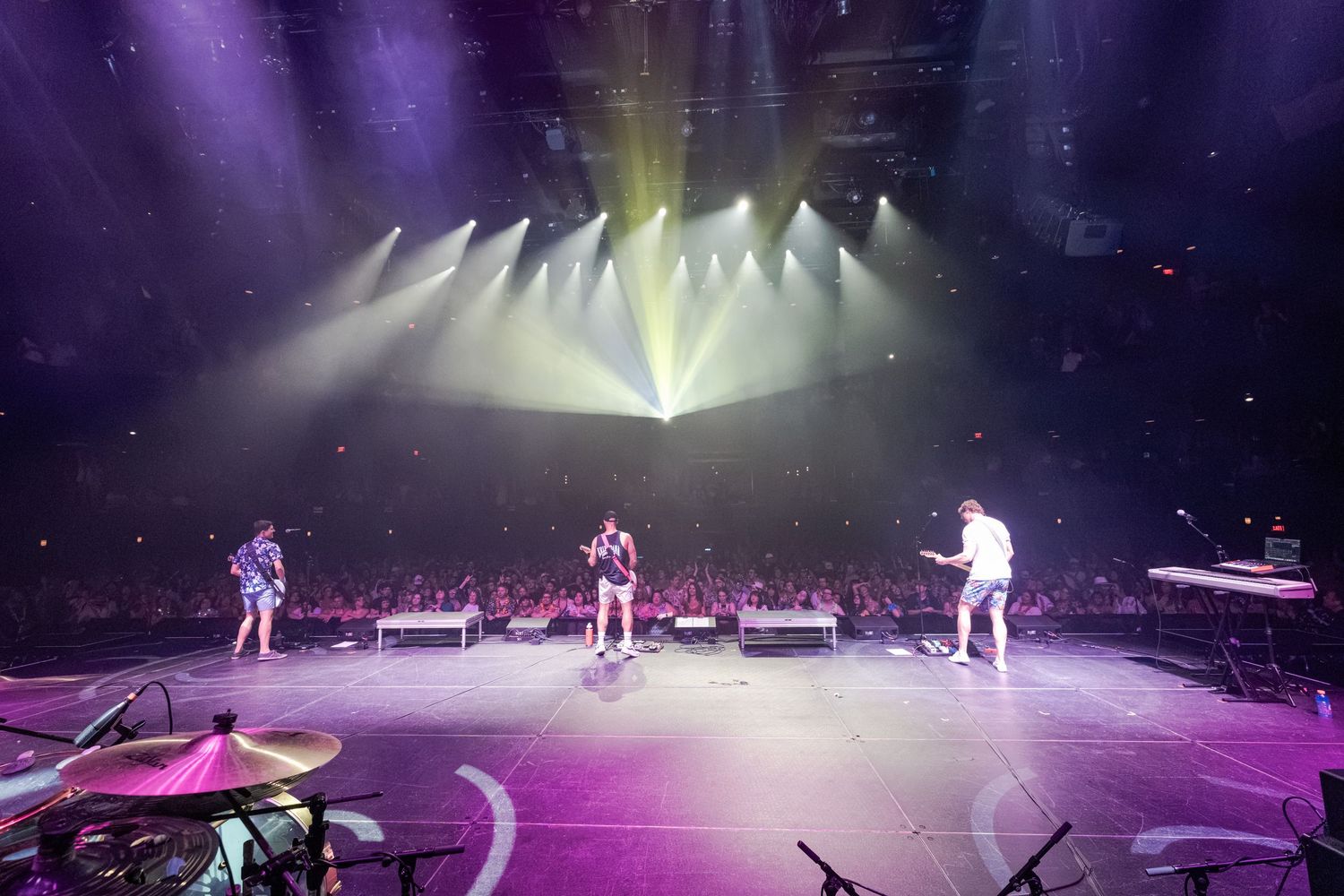 Three performers on a round stage with dramatic purple and white lighting at a packed concert arena.