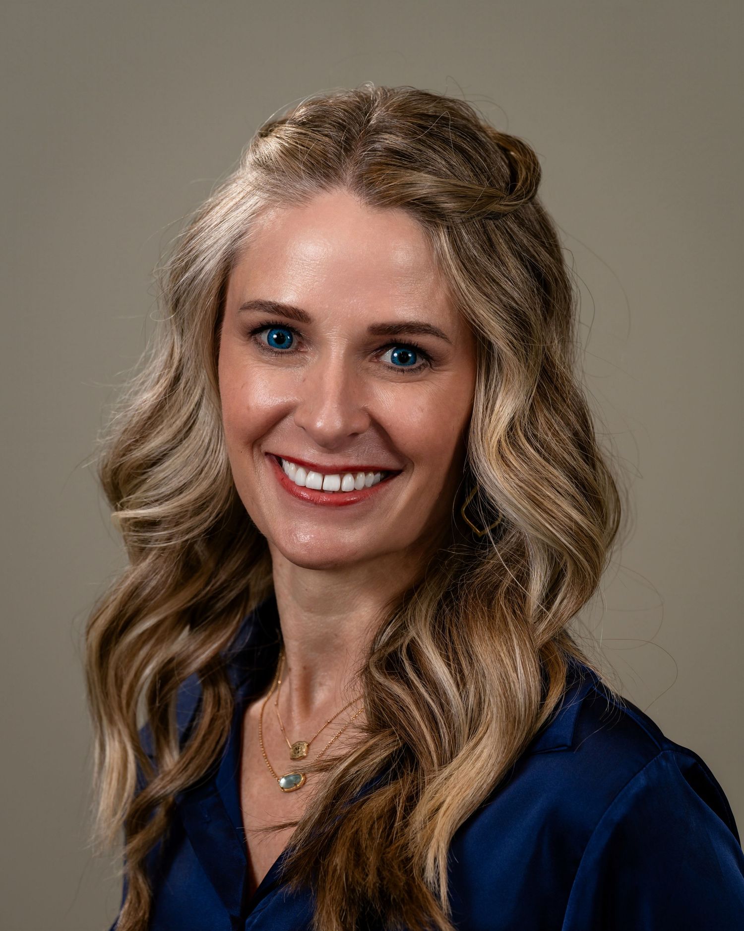 Professional headshot of a smiling blonde woman with blue eyes, wearing a navy blouse and gold necklace.