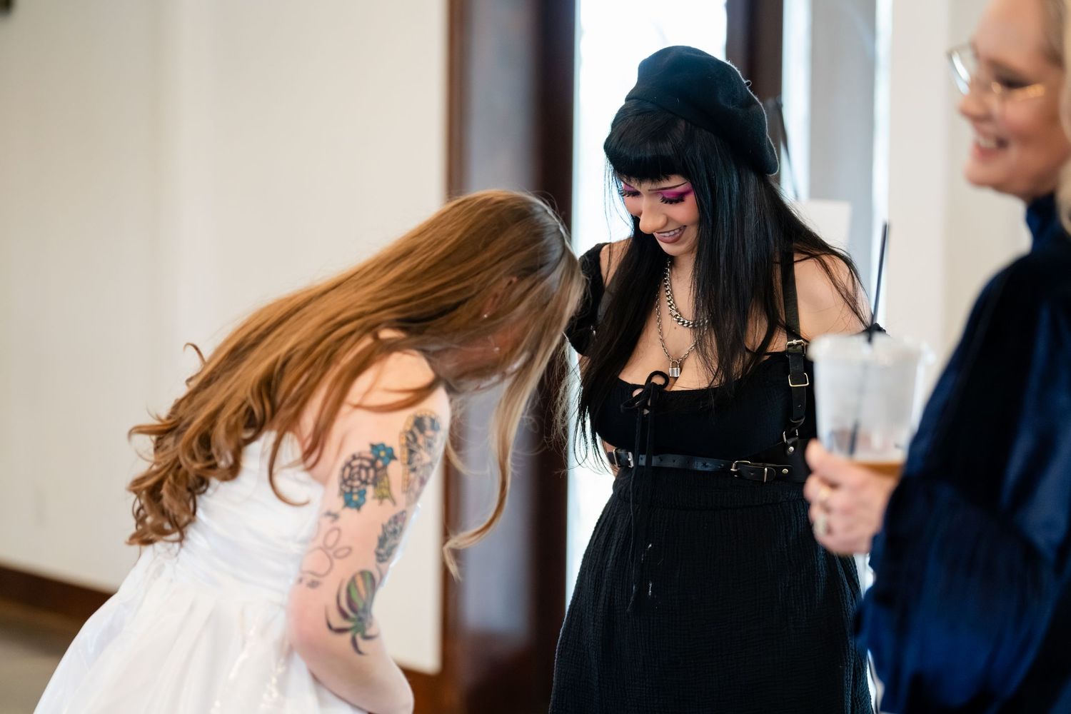 Two women at a party, one in white dress with tattoos, another in black gothic outfit with beret, sharing a joyful moment.