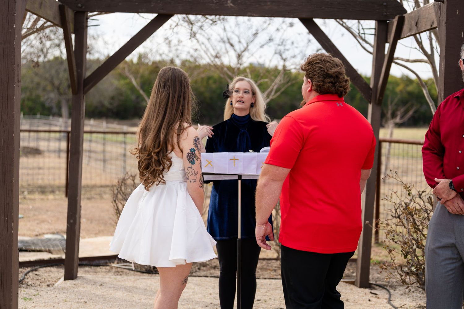 Outdoor wedding ceremony under a wooden pergola with officiant and couple in a rustic farm setting.