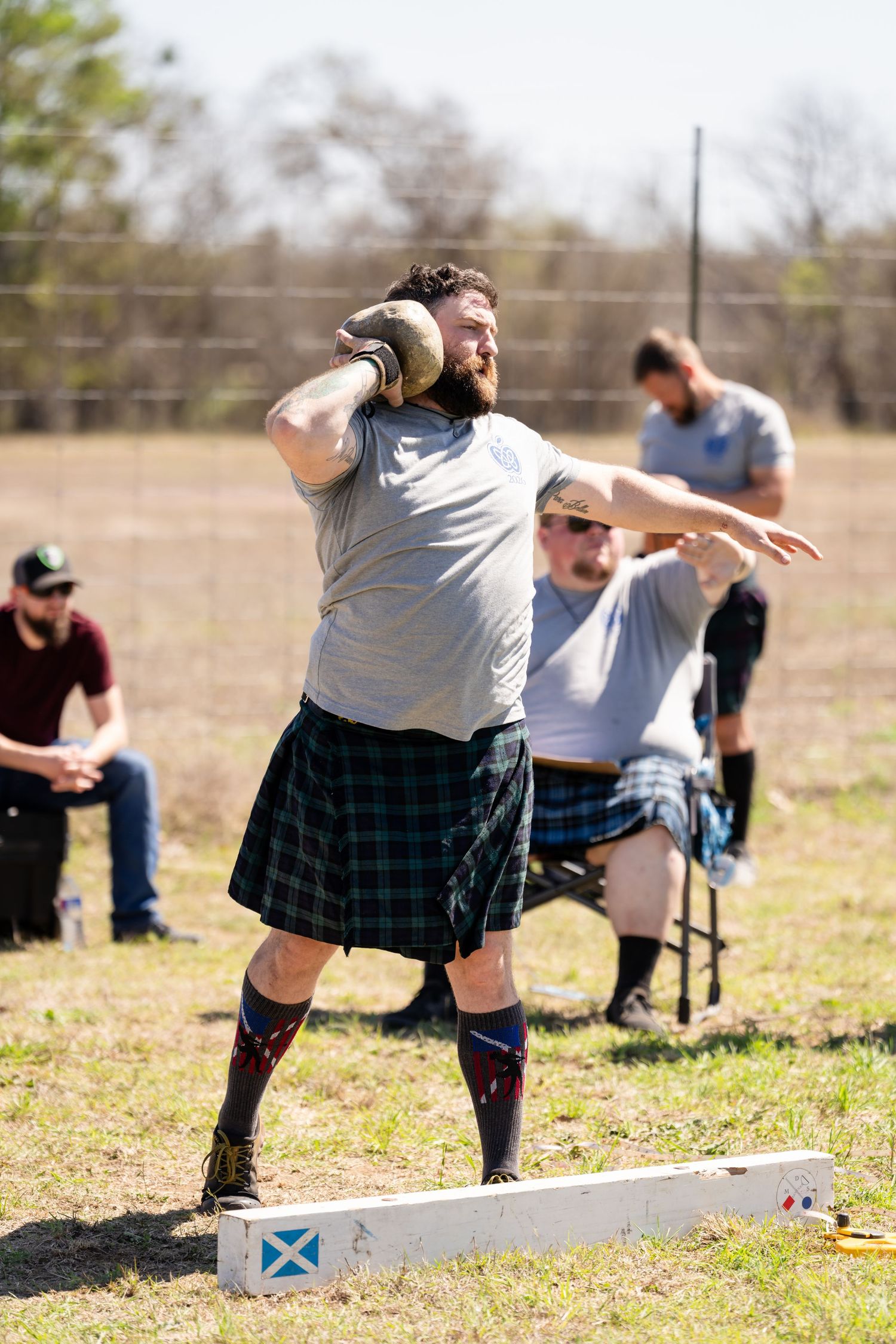 Man in tartan kilt performs shot put throw at Highland Games competition outdoors.