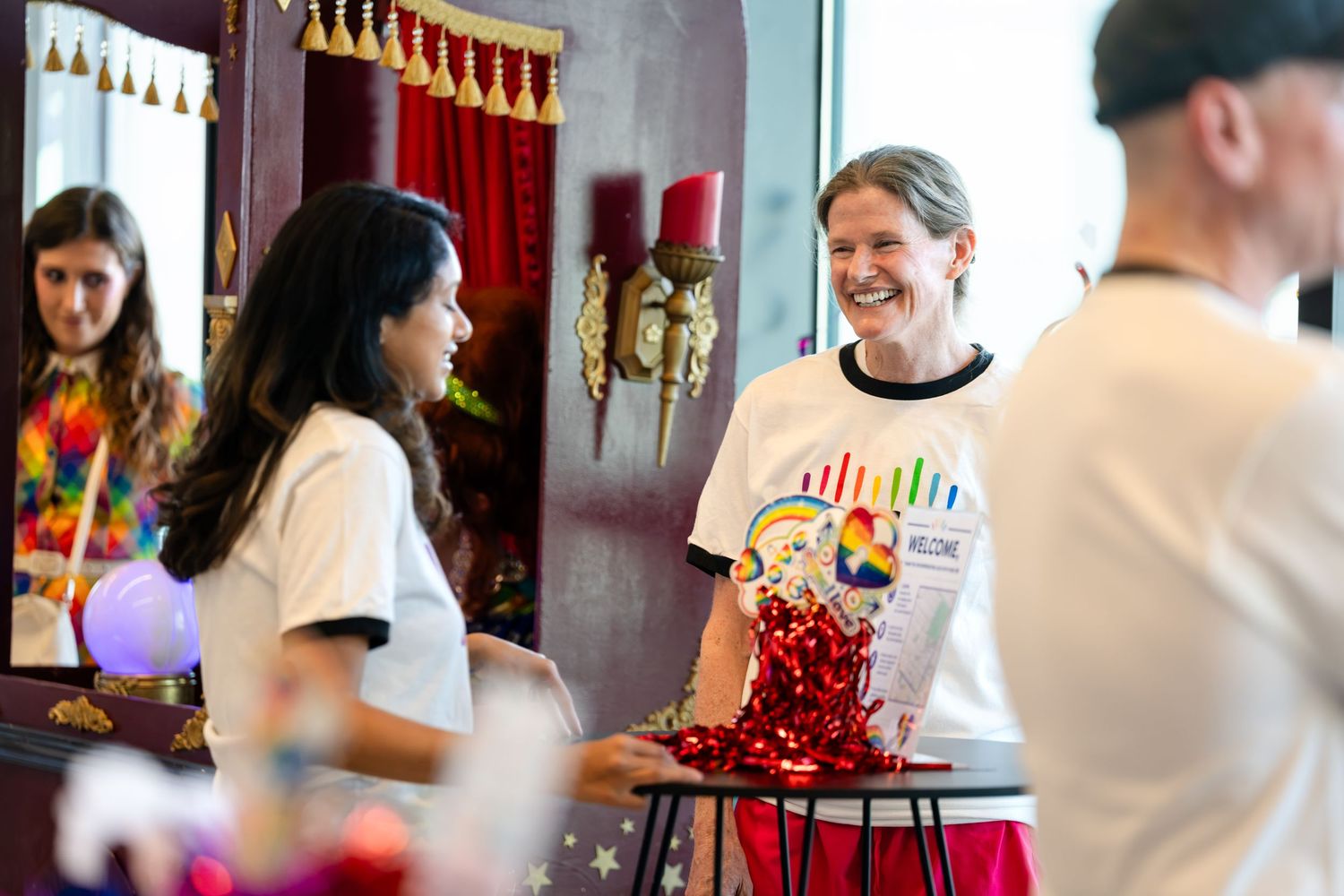 People in matching white t-shirts smile and interact at a colorful Pride event indoors.