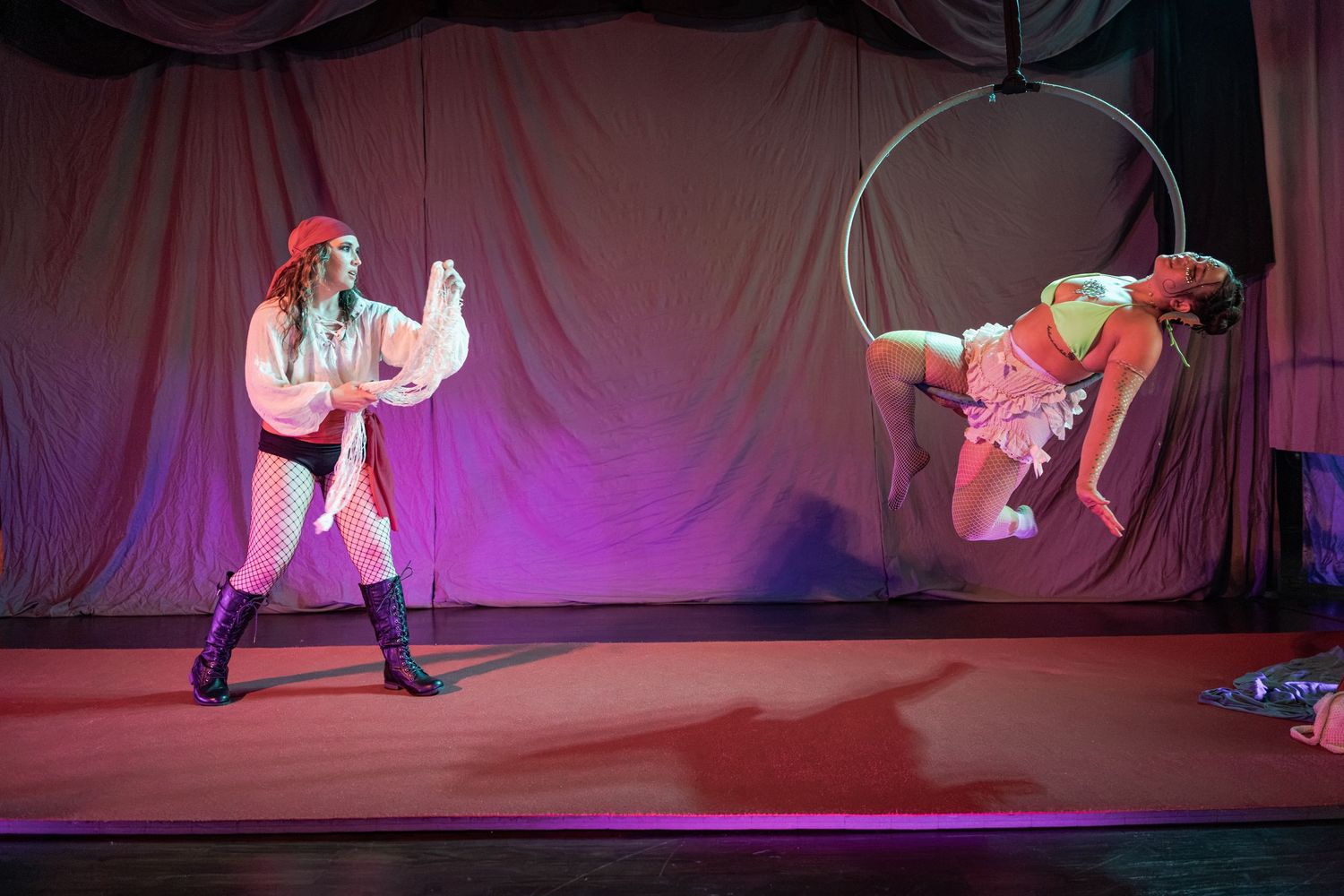 Circus performers on stage, one in pirate costume and another doing aerial acrobatics on a hoop under purple lights.