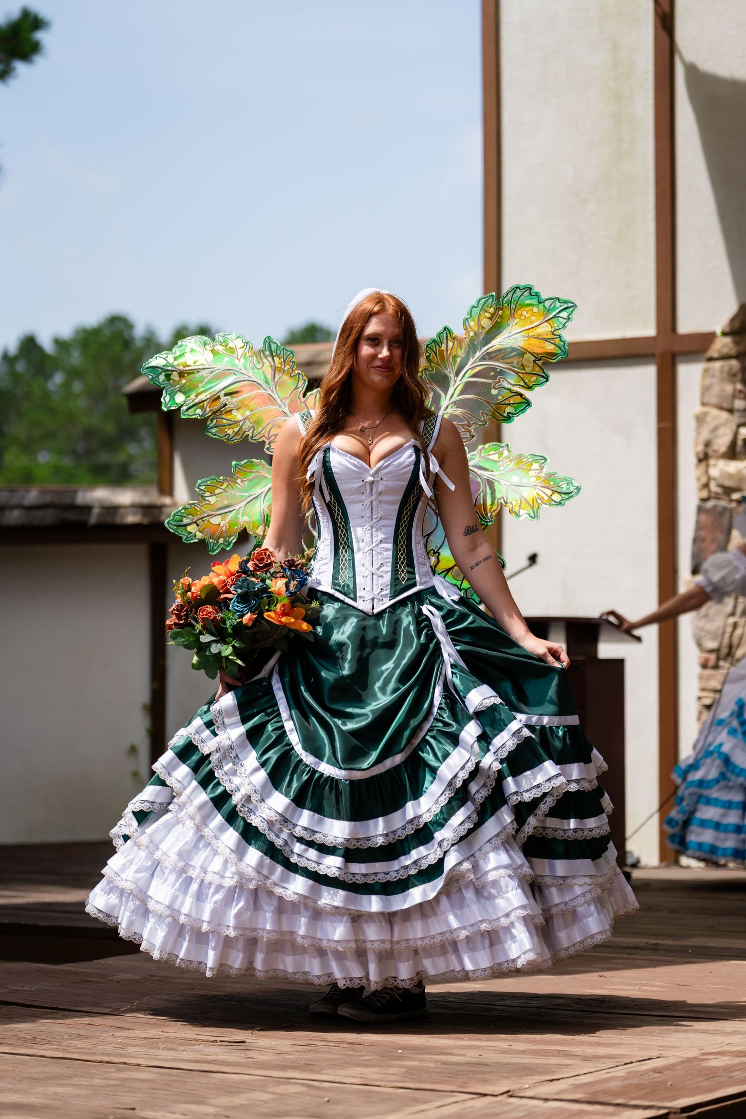 Woman in green fairy costume with wings and flower bouquet performing on outdoor stage at Renaissance fair.