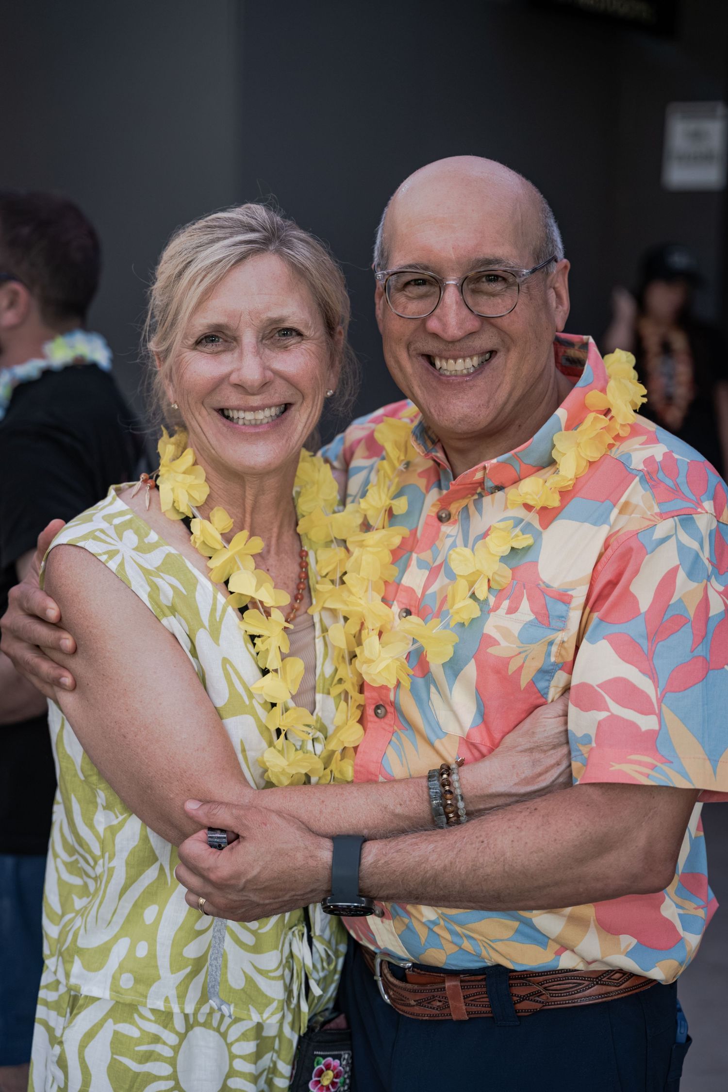 Smiling couple wearing yellow leis and Hawaiian attire pose together at a tropical-themed party event.