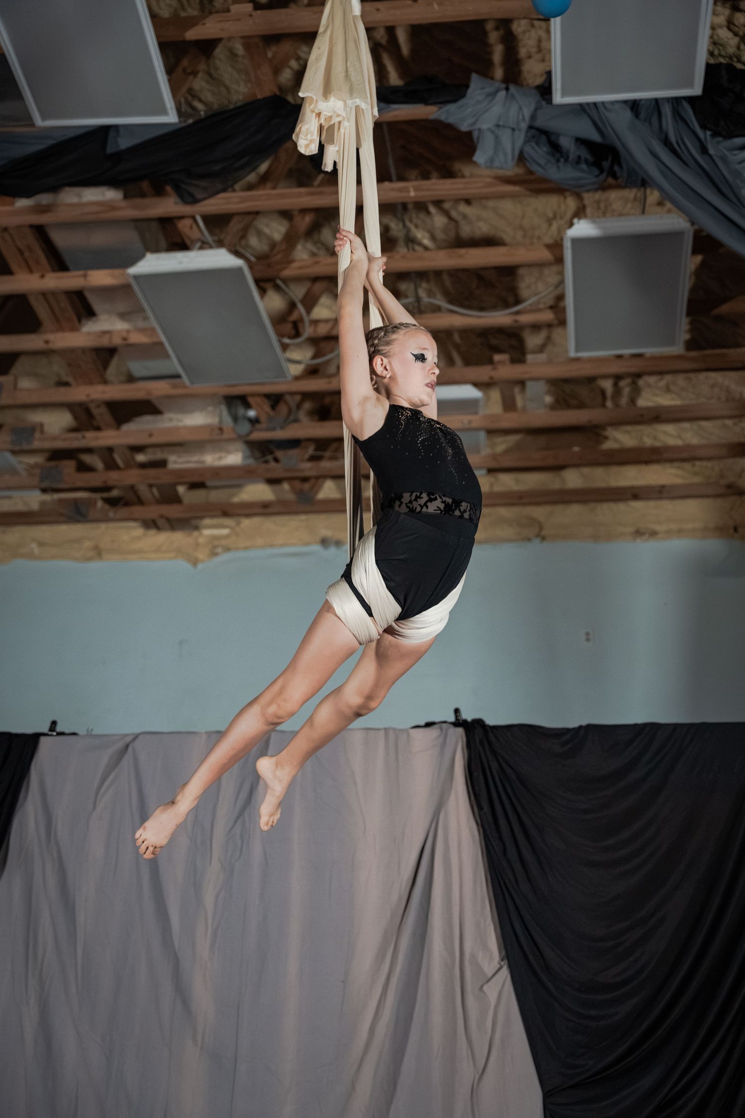Young aerial silk performer in black leotard suspended mid-air during practice in rustic indoor studio.