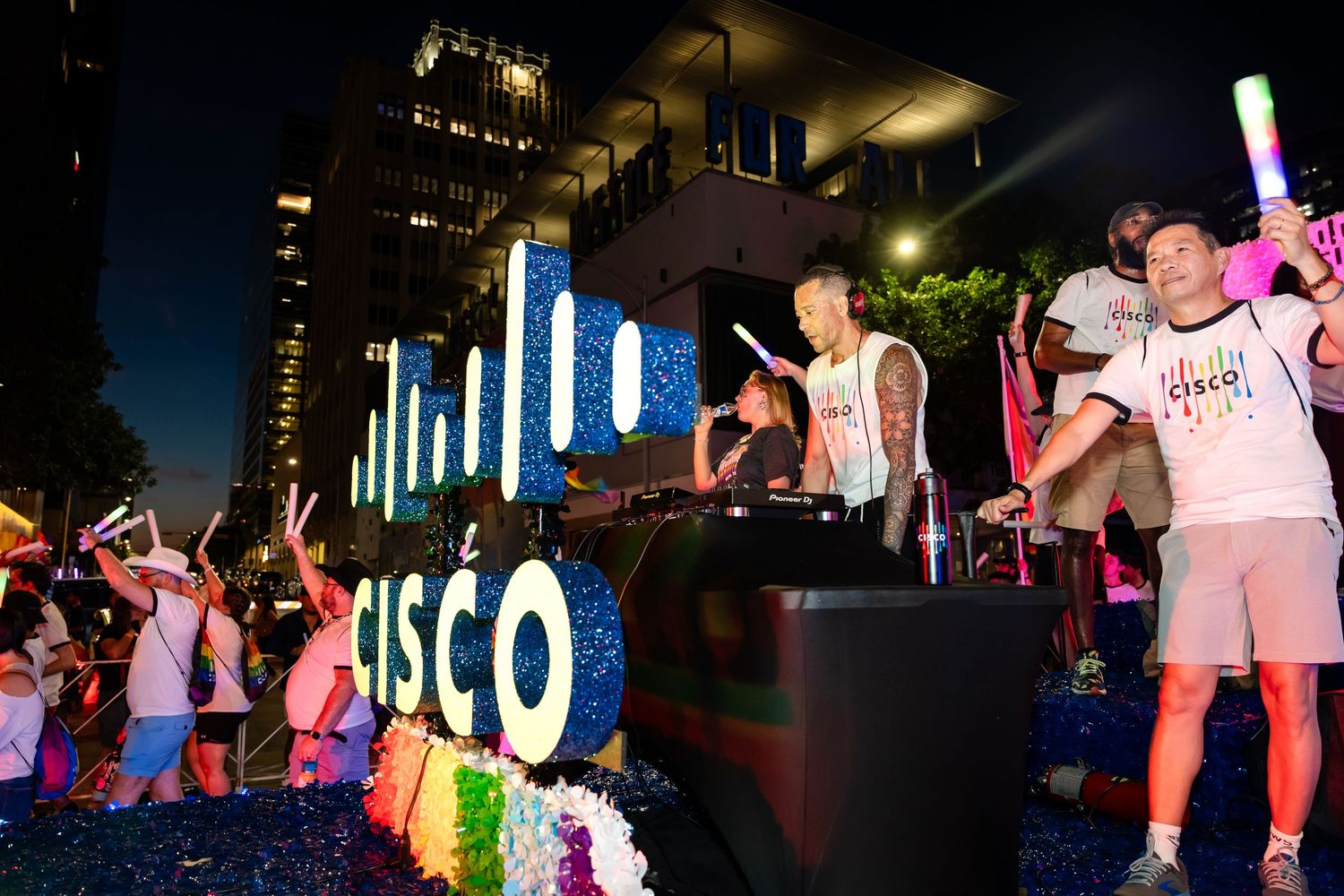 People celebrate at a colorful Cisco Pride parade float at night with glowing lights and festive decorations.