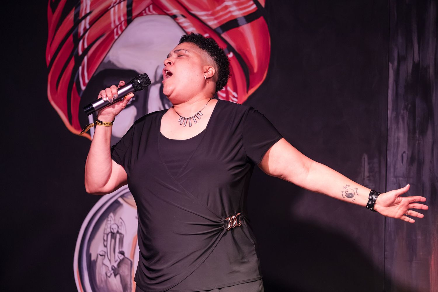 Performer in black t-shirt sings passionately on stage with dramatic red lighting and dark background.