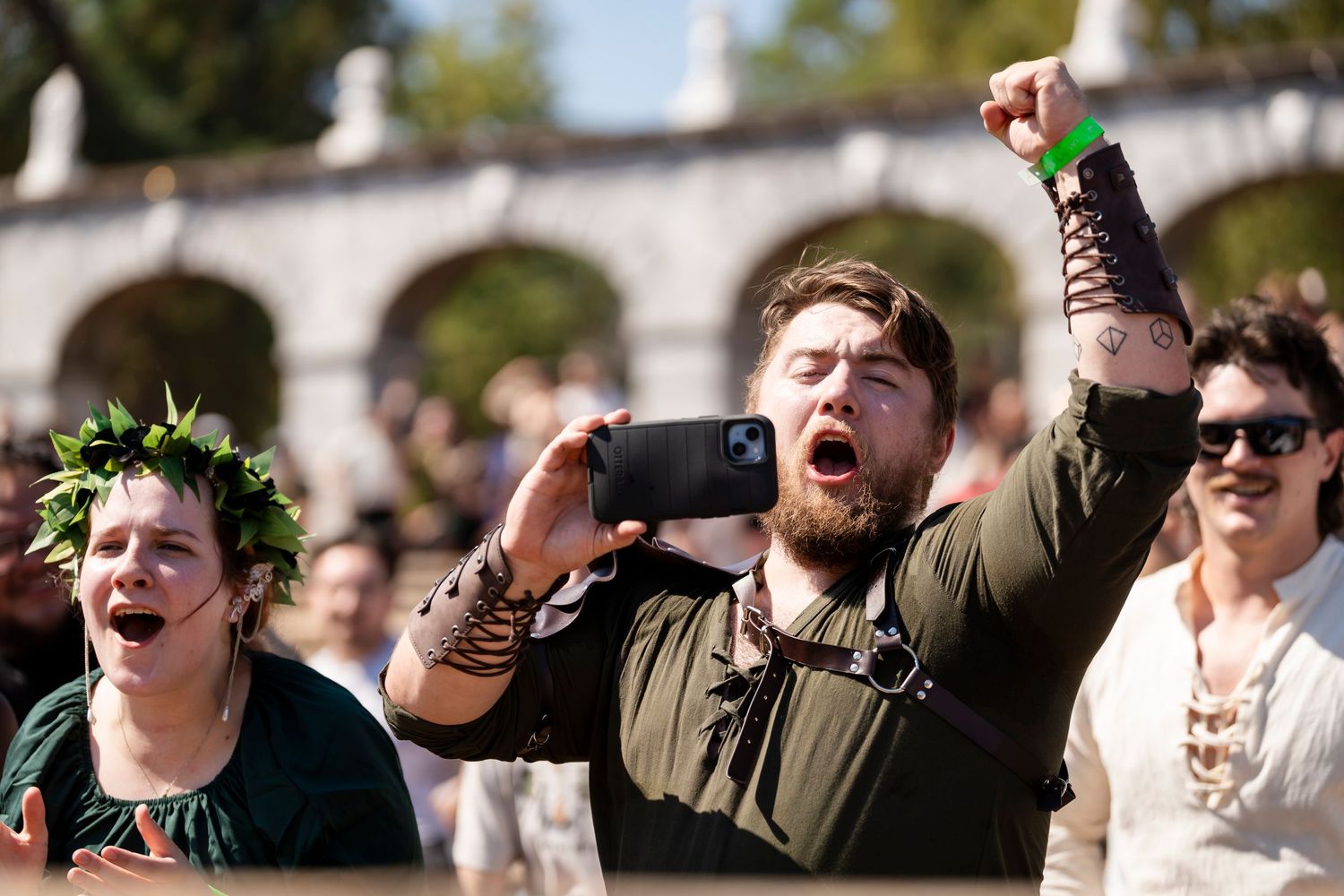 Excited festival attendees wearing green laurel wreaths cheer and raise their drinks at an outdoor celebration.