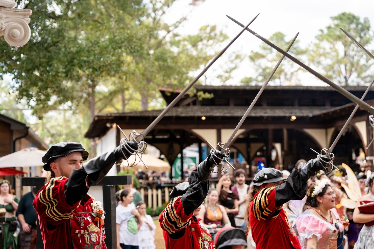 Three performers in red Renaissance costumes demonstrate sword fighting techniques at an outdoor festival.