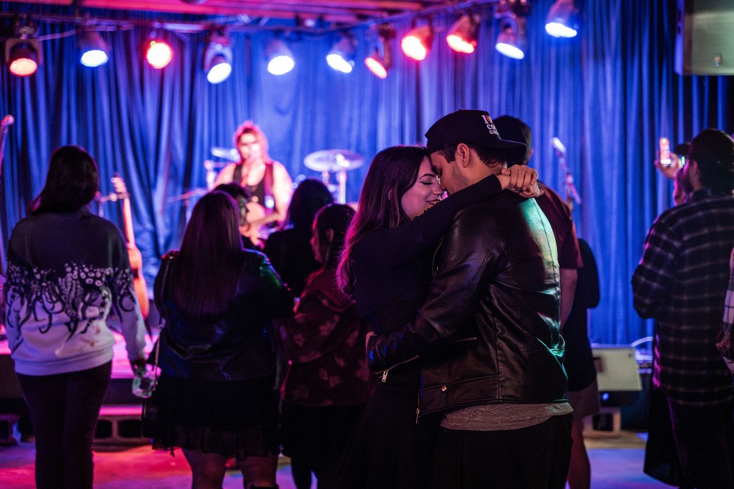 People dancing and socializing at a nightclub with blue curtains and colorful stage lighting overhead.