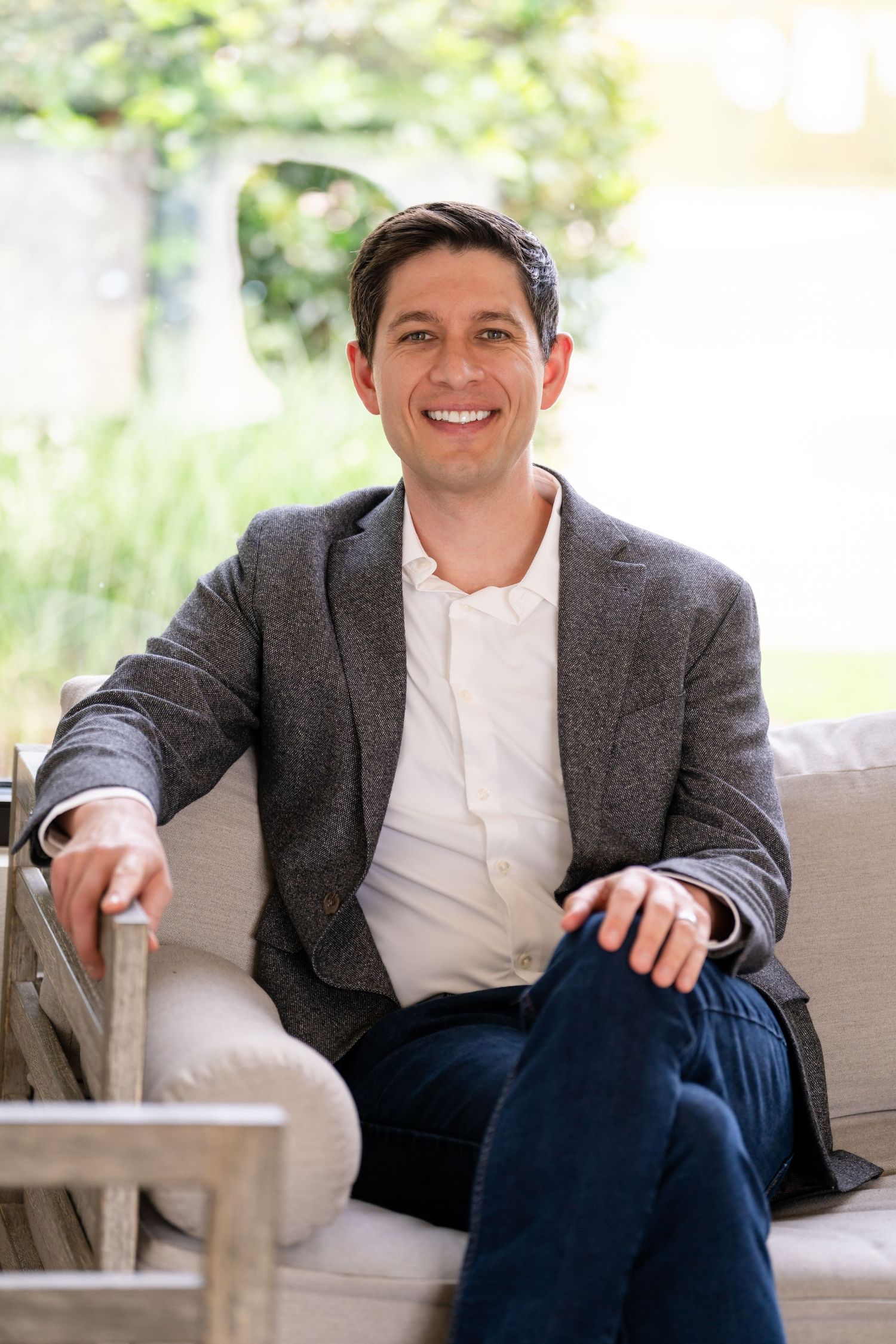 Smiling man in gray blazer and white shirt sitting relaxed on a beige sofa outdoors.