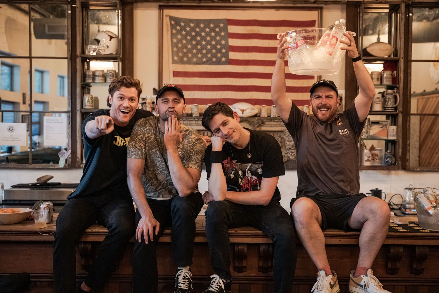 Four friends pose happily in front of an American flag display inside a rustic bar or restaurant setting.