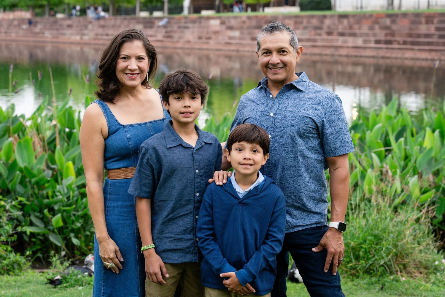 Family of four smiling outdoors near a pond, parents and two boys wearing coordinating blue outfits in a park setting.