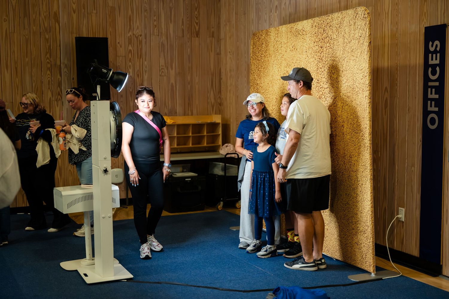 People gathered indoors near a photo booth setup with wooden panel backdrop in a modern office space.