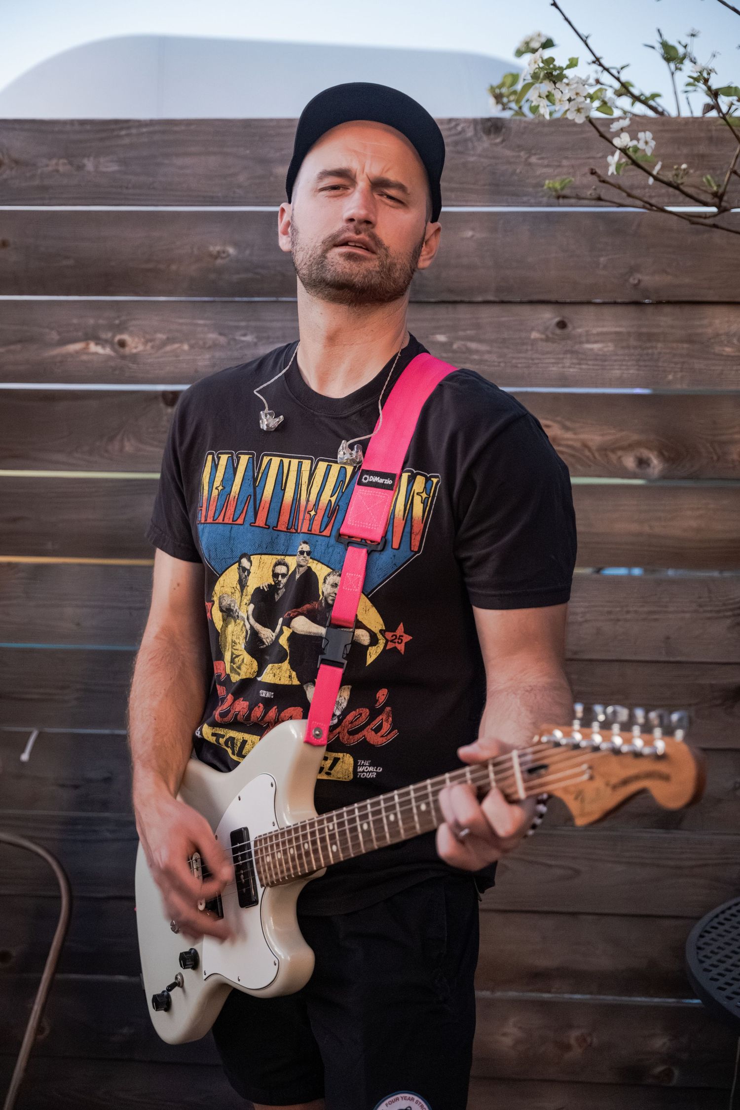Man in black cap and band tee plays white electric guitar with pink strap outdoors against wooden fence.