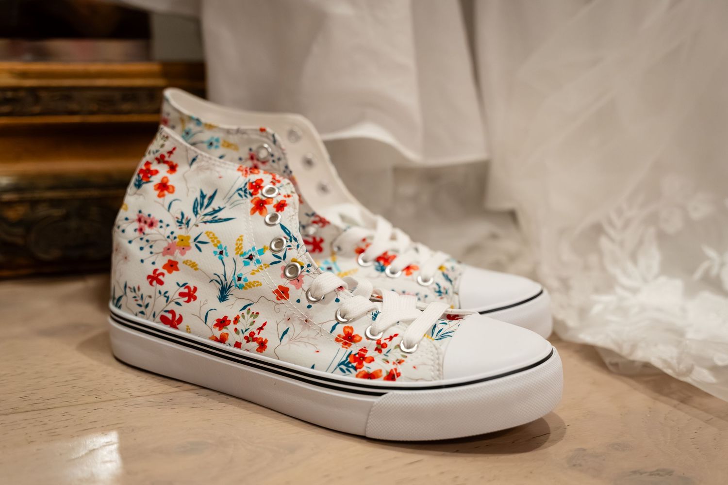 White high-top sneakers with colorful floral pattern displayed near a wedding dress on a wooden floor.