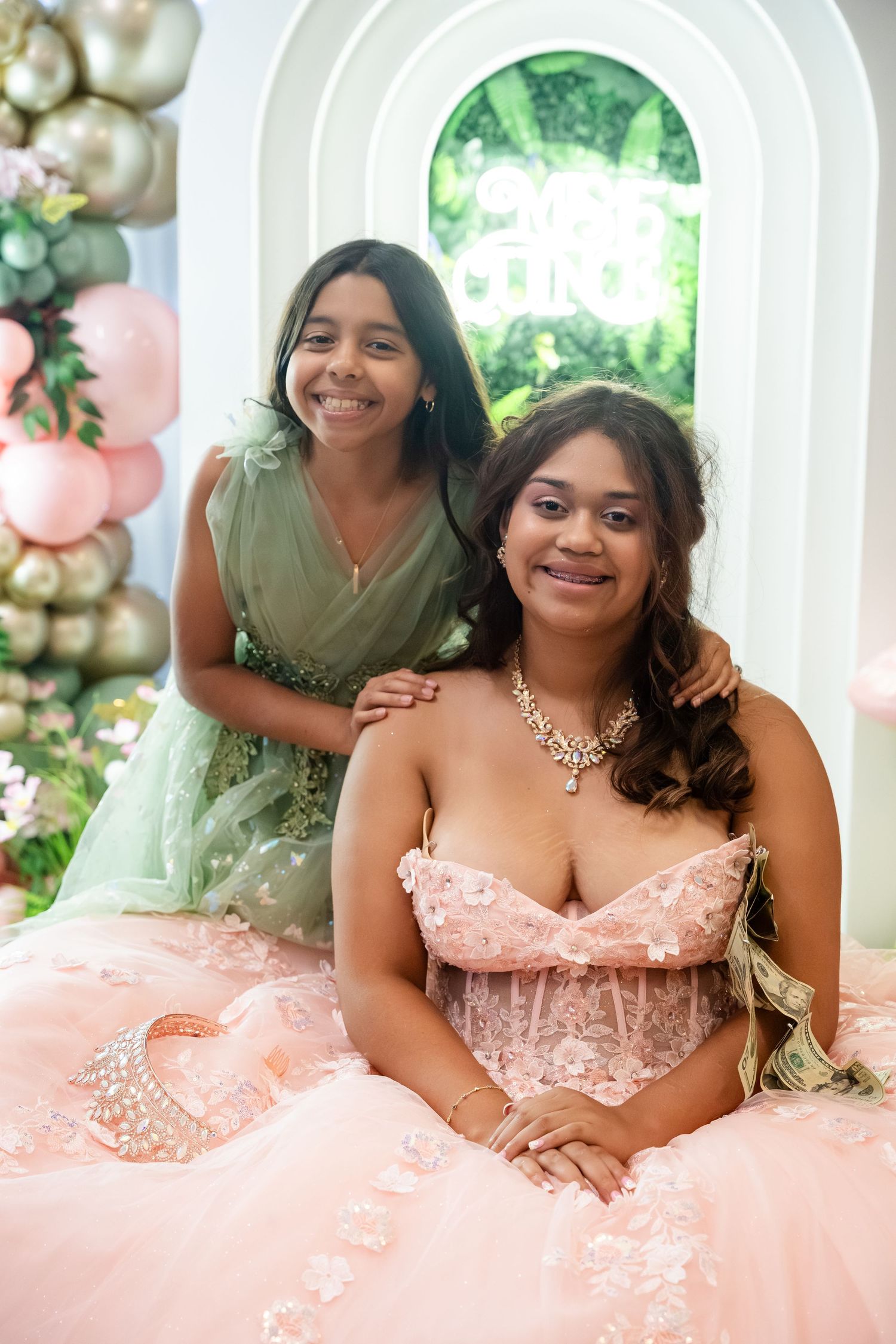 Two sisters posing at a quinceañera, one in a pink ball gown with tiara and one in a green dress.