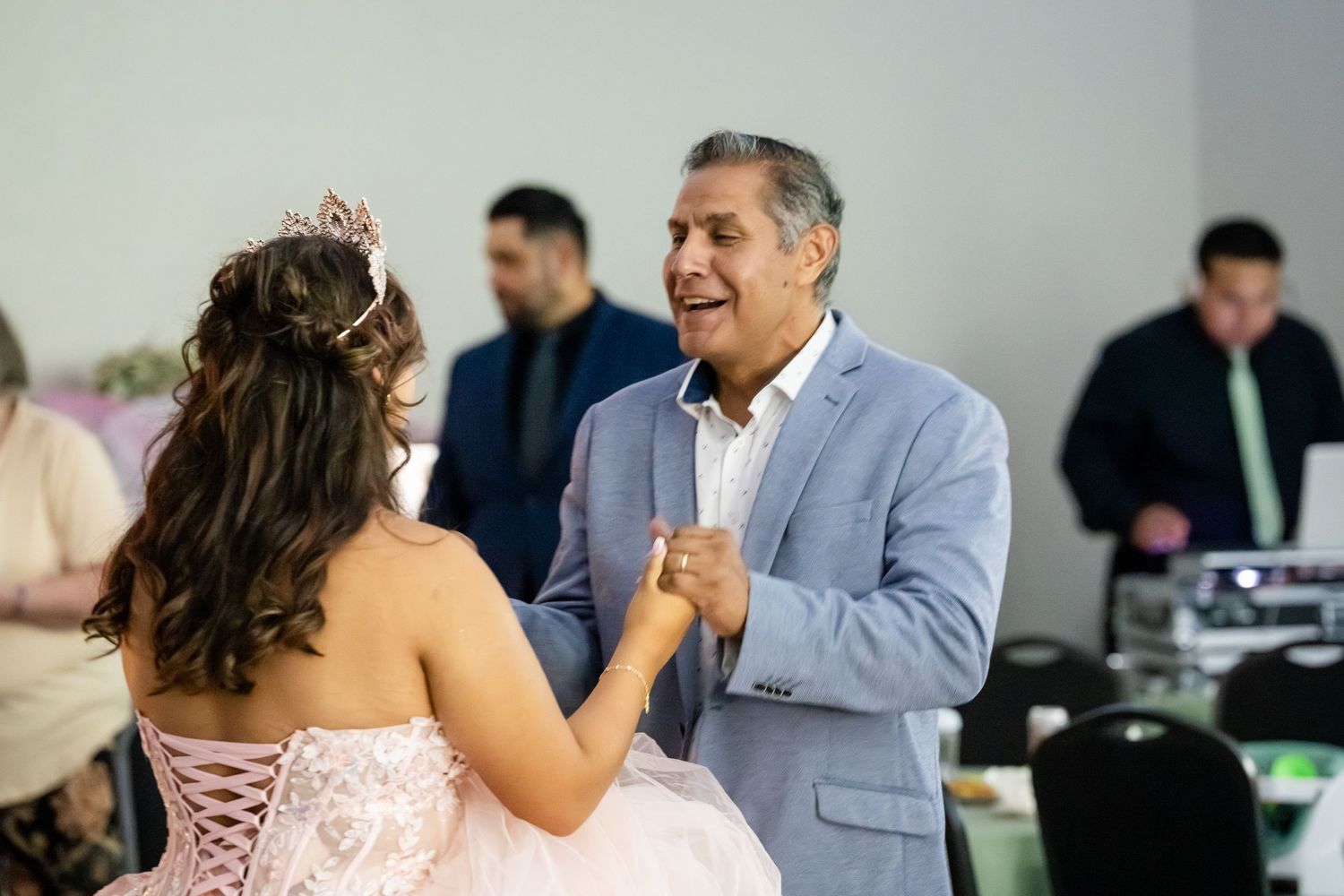 A man in a blue suit dances with a young woman in a pink quinceañera gown and tiara at a celebration.