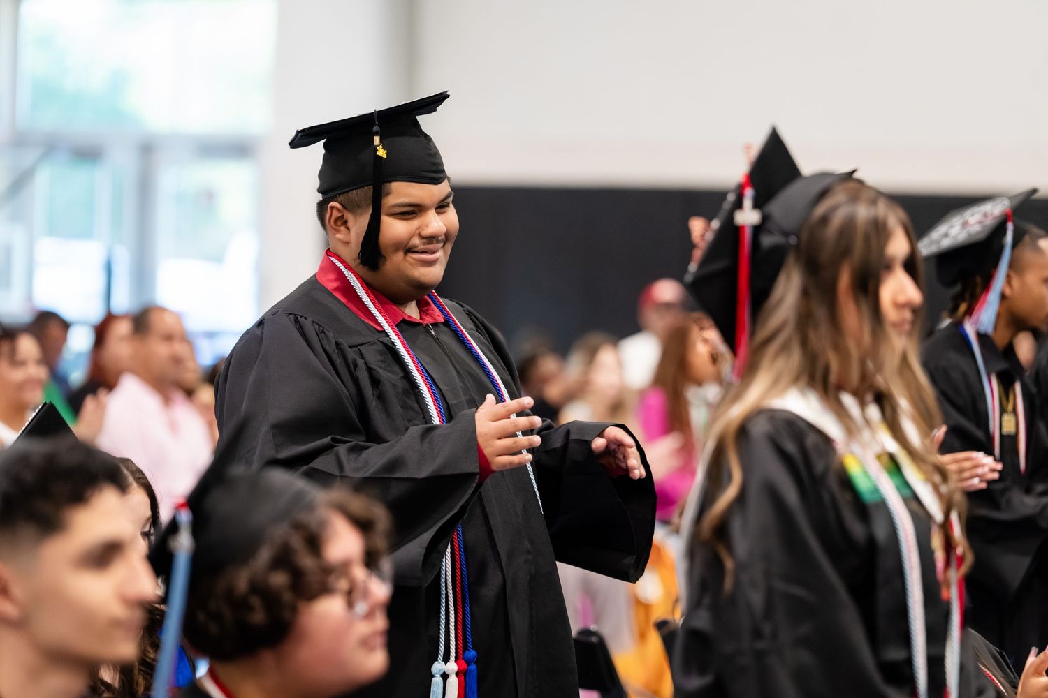 Smiling graduate in black cap and gown walks during graduation ceremony among fellow graduates.
