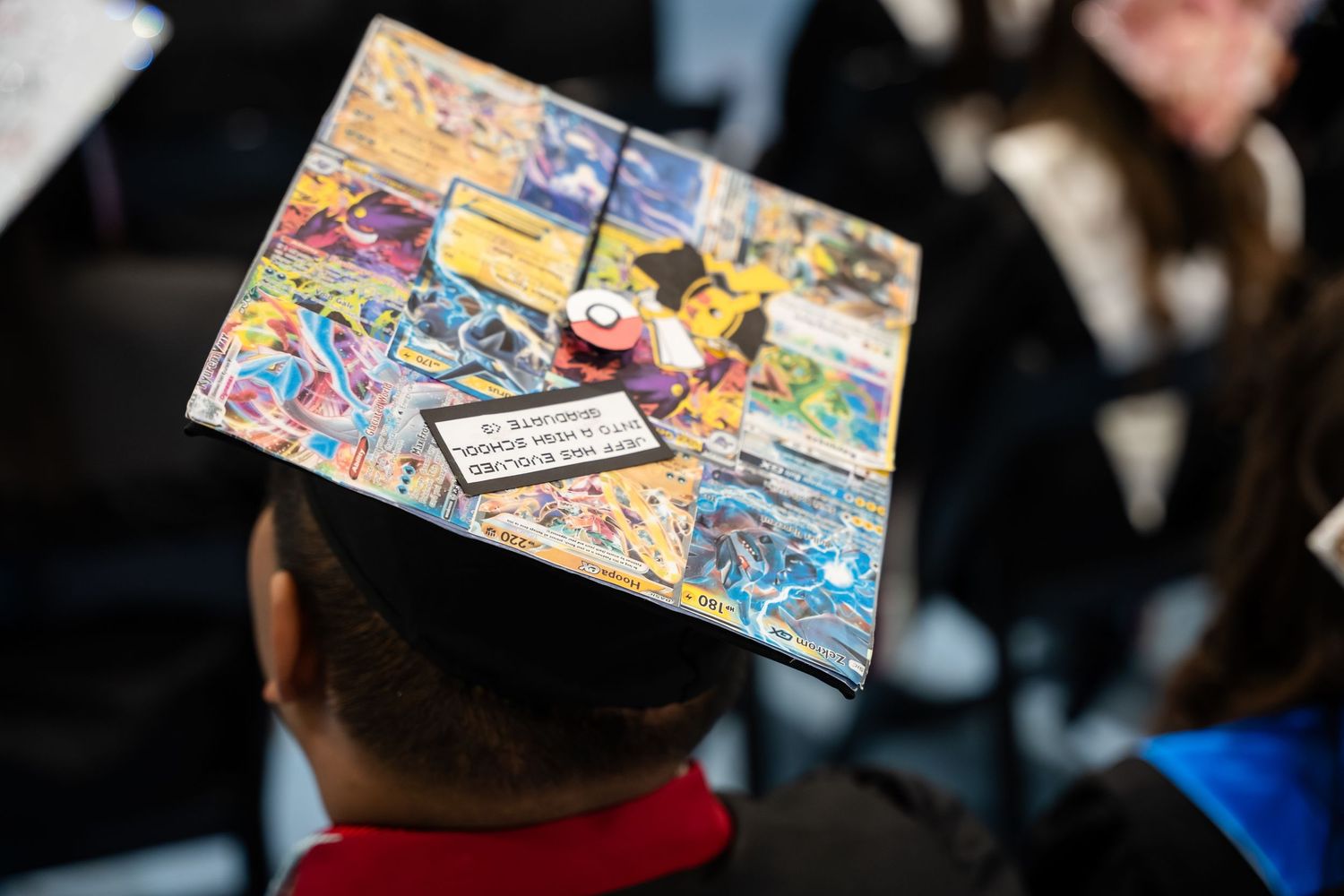Graduate wearing a Pokemon trading card decorated mortarboard cap at a graduation ceremony.