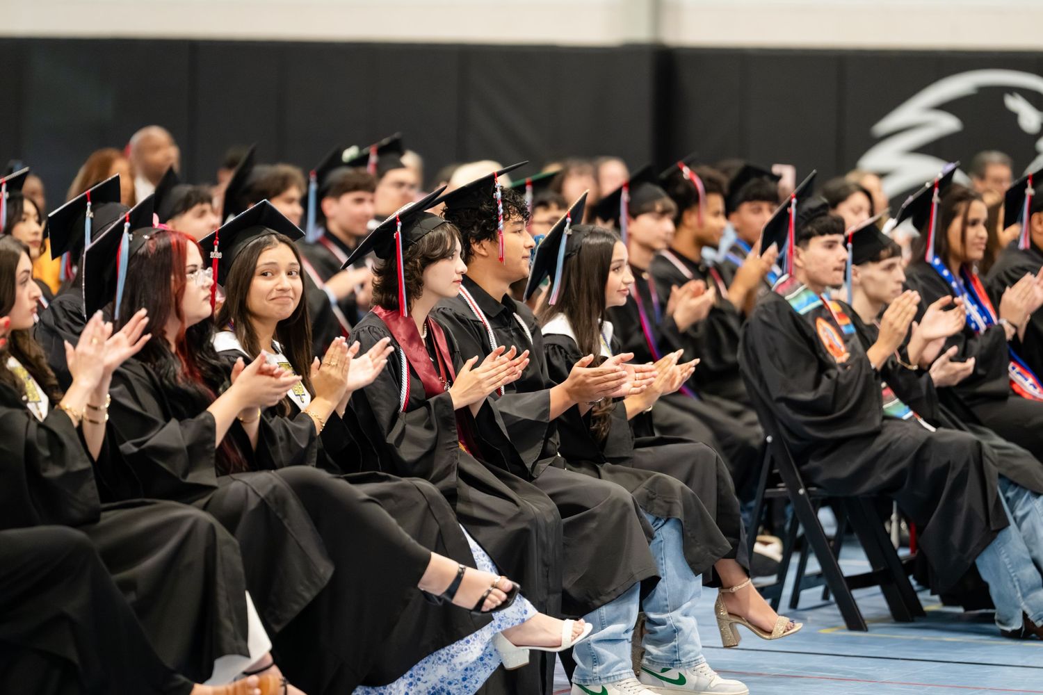 Graduates in black caps and gowns clapping during a commencement ceremony seated in a gymnasium.