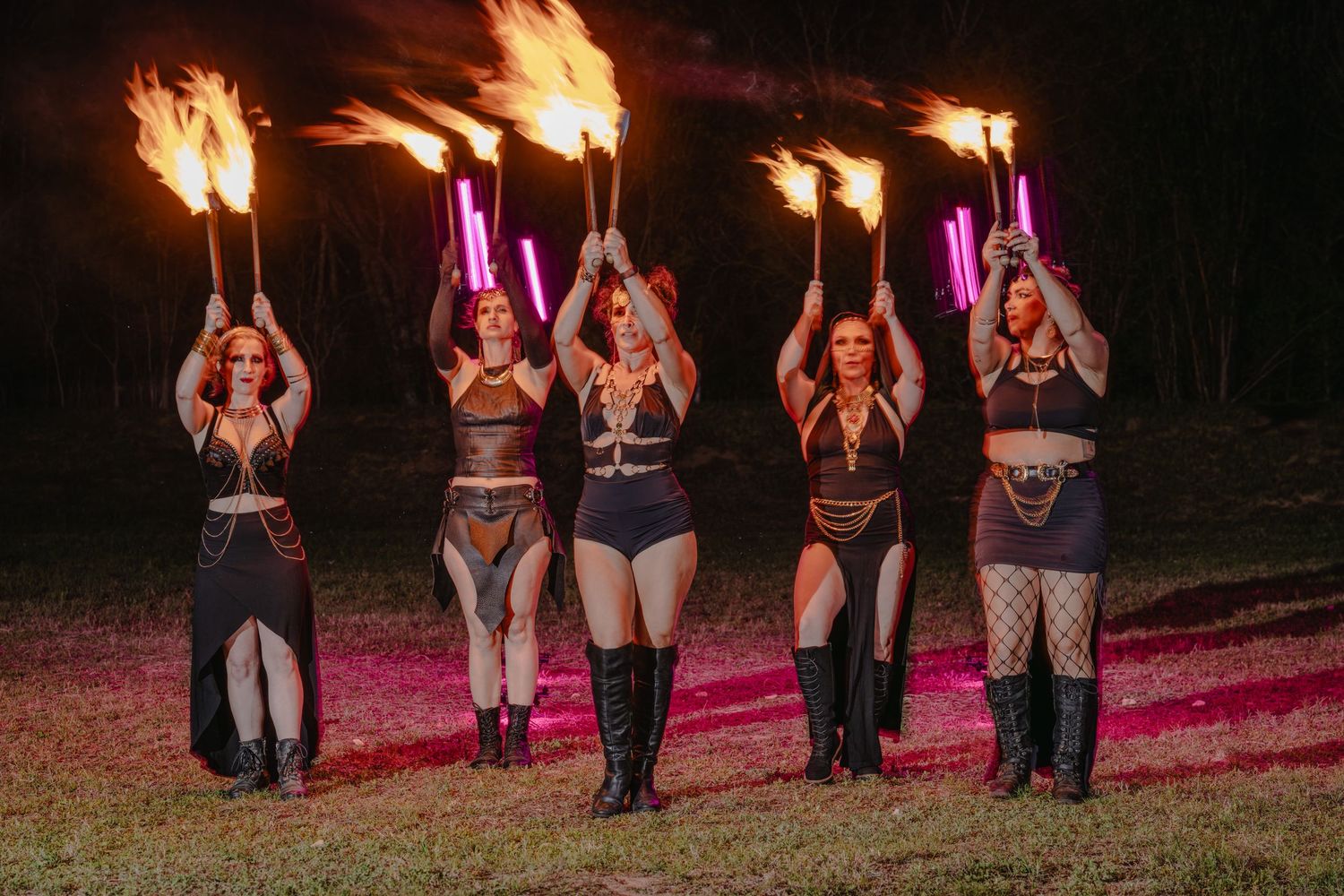 Five female fire performers holding flaming torches and glowing pink LED props during an outdoor night show.