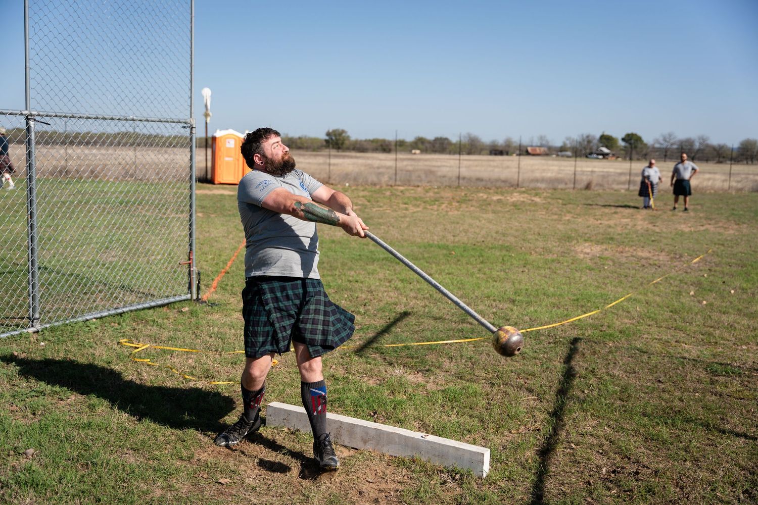 Man in tartan kilt throwing hammer at Highland Games competition on grassy field outdoors.