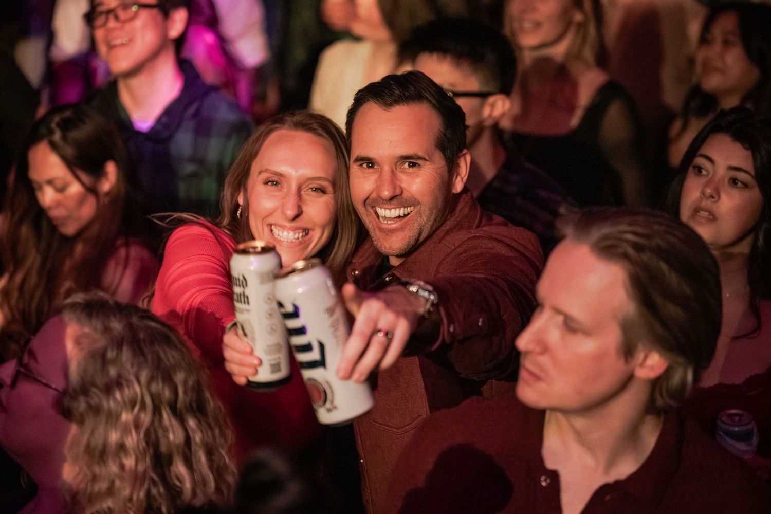 Two smiling friends holding Miller Lite beer cans at a lively crowded party event.